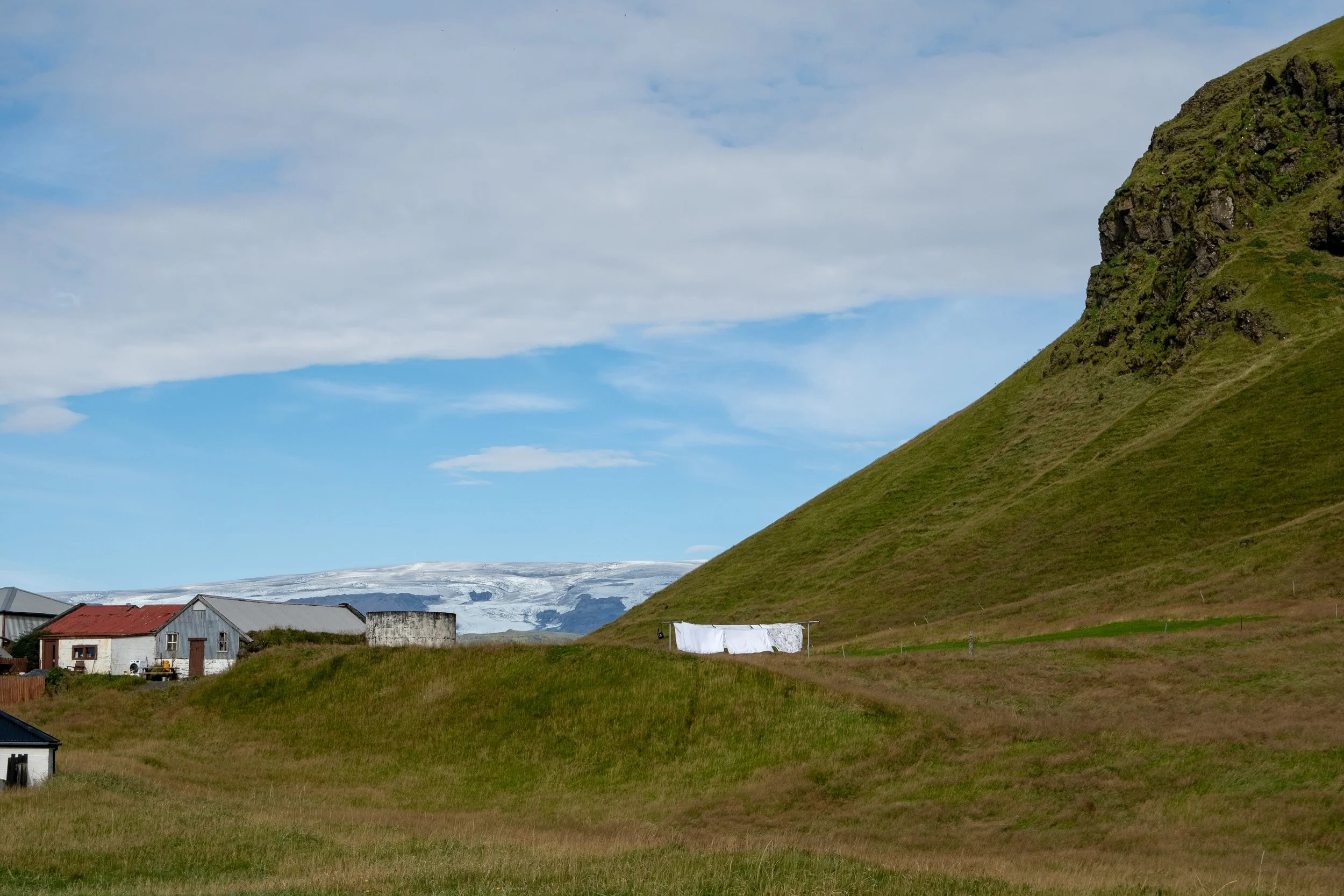 A rural landscape with grassy hills, a mountain on the right, small houses with metal roofs, and a snow-capped glacier in the background under a partly cloudy sky.