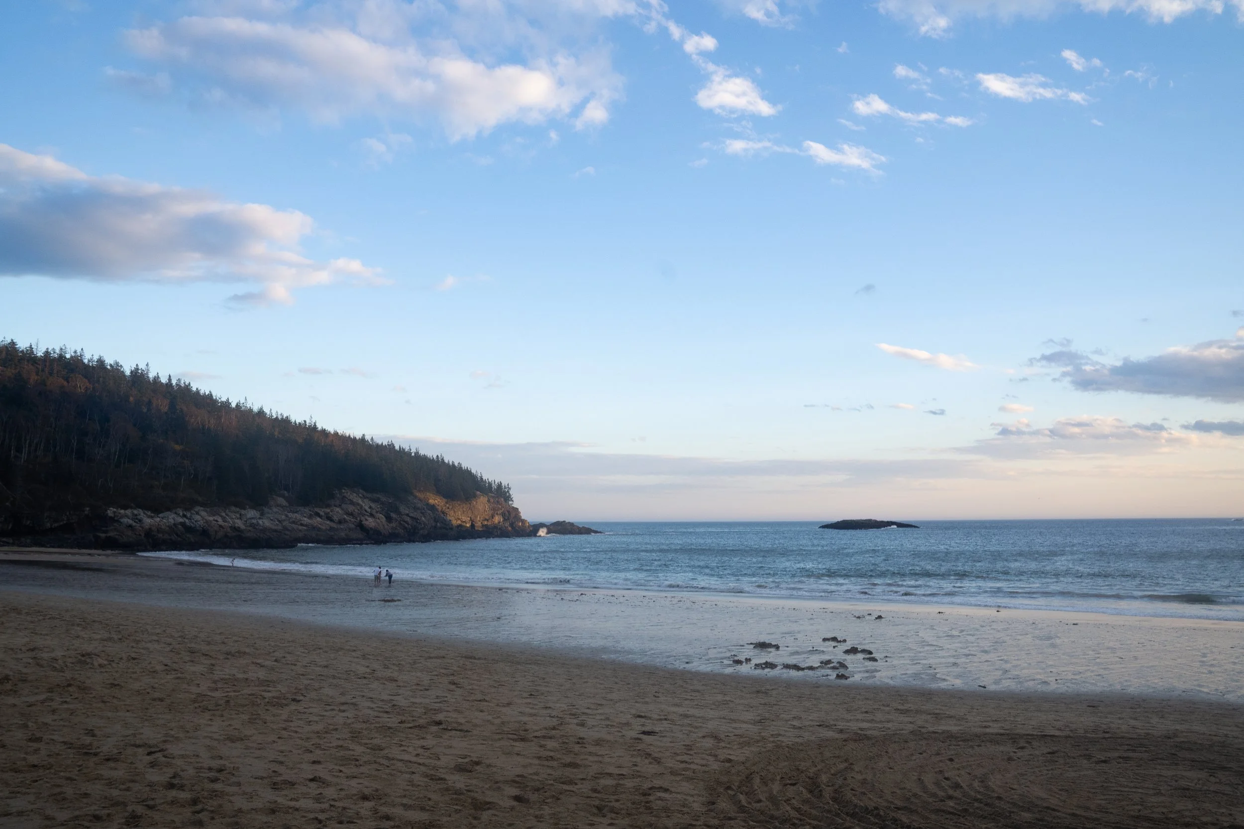 A beach scene with sandy shore, calm ocean water, and a forested hill in the background under a partly cloudy sky.