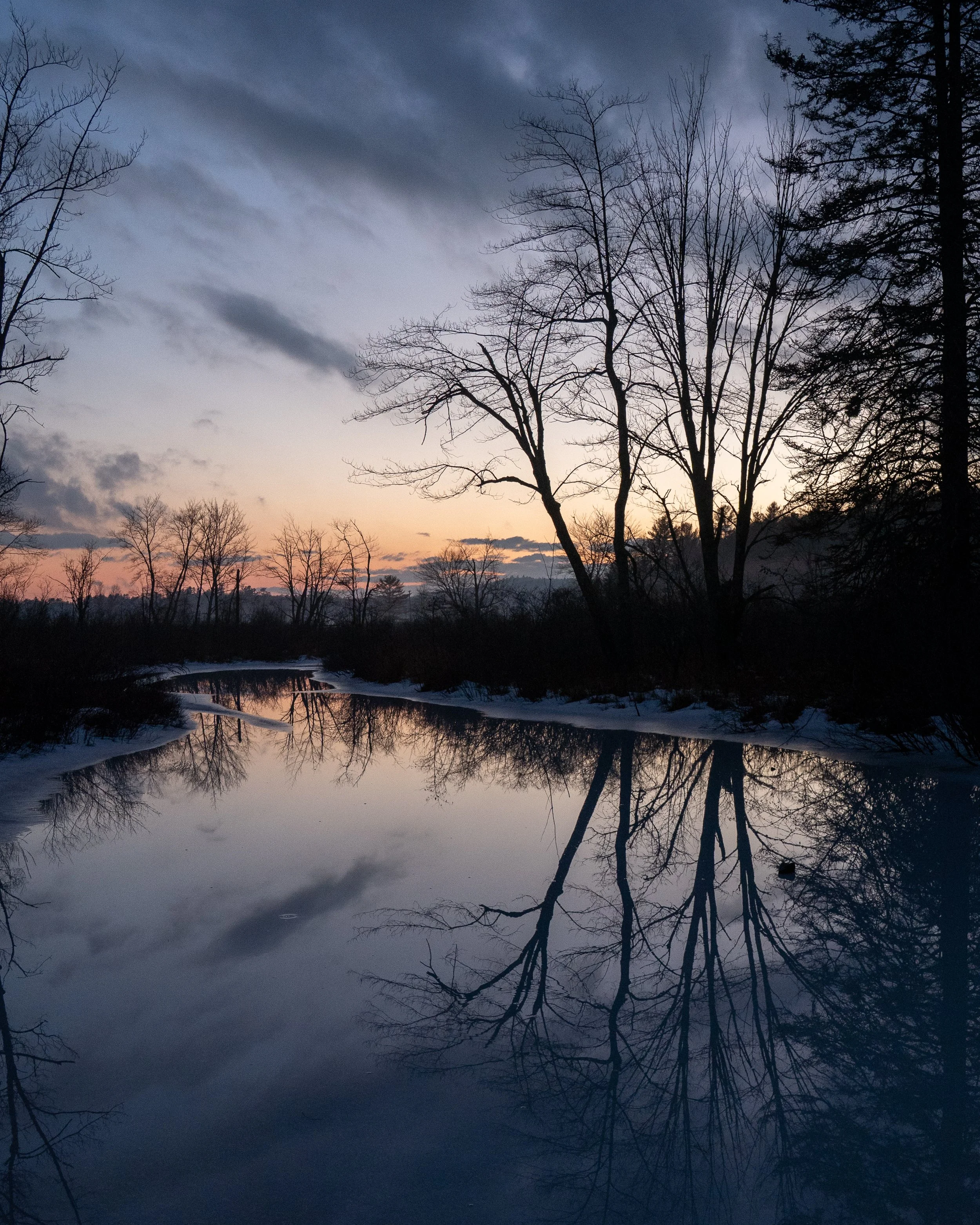 A peaceful river scene during sunset with leafless trees reflecting in the calm water, and dark clouds overhead.