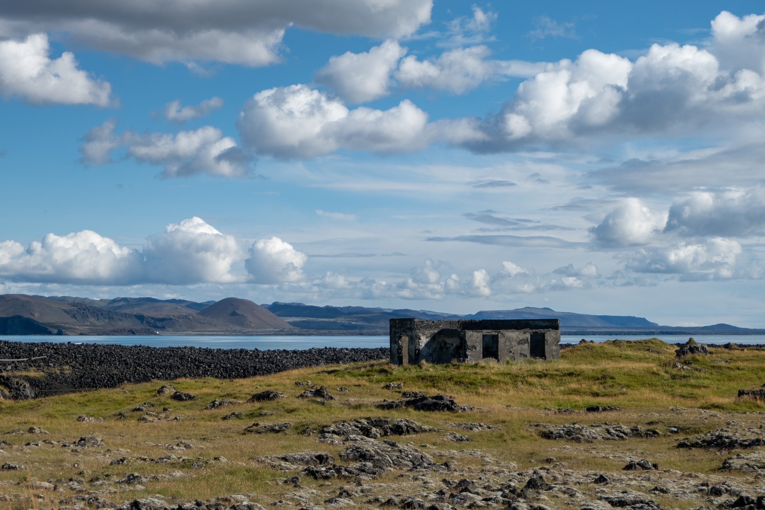 A grassy landscape with rocky ground, a small stone building, a body of water, distant mountains, and a partly cloudy sky.