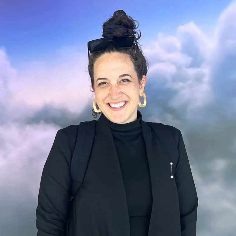A young woman with curly brown hair tied up, smiling, wearing a black turtleneck, black jacket, large gold hoop earrings, and sunglasses on her head, standing in front of a screen with a purple cloudy sky background.