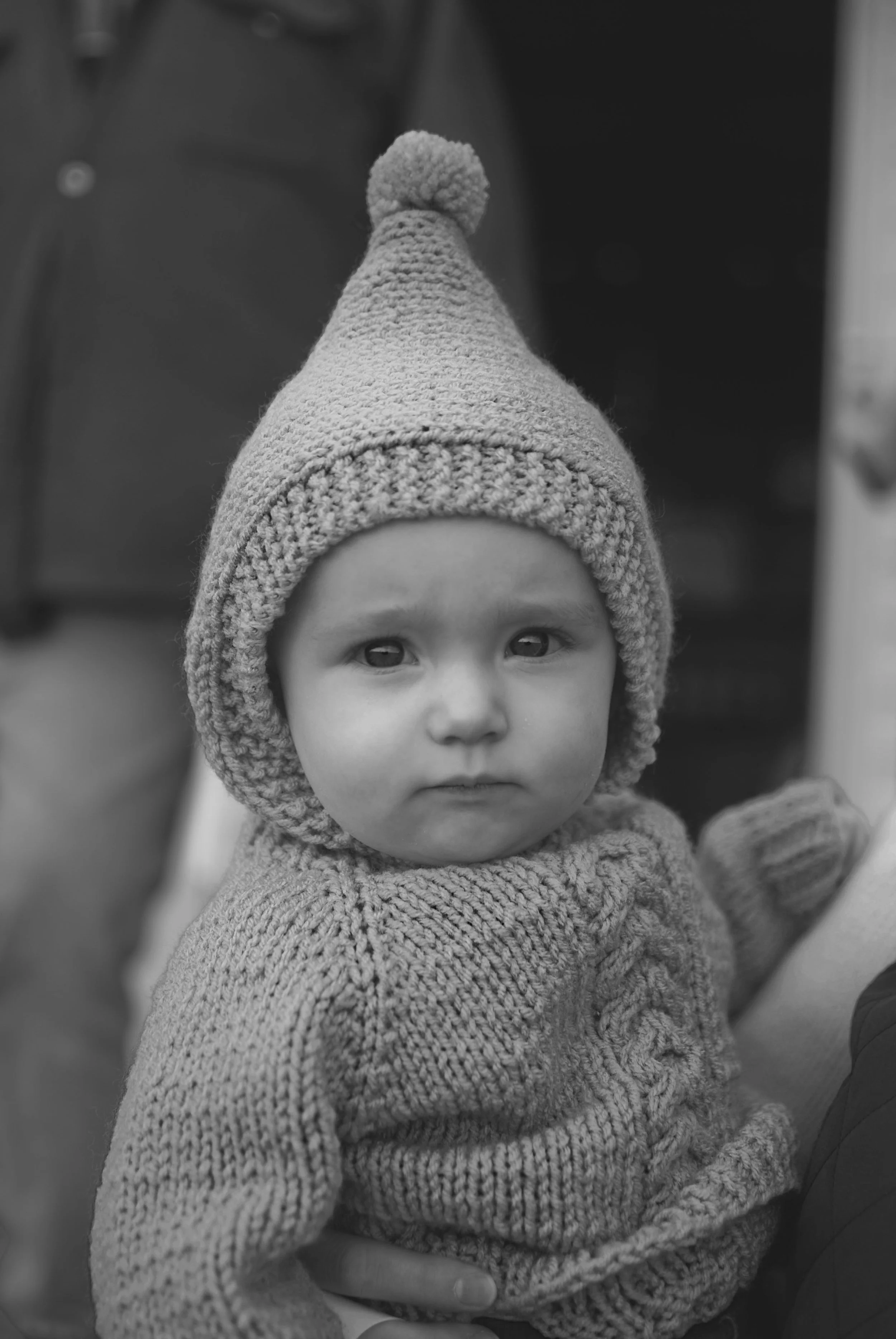 A young child wearing a cozy knit hat with a pom-pom on top and a knit sweater, looking directly at the camera in black and white.