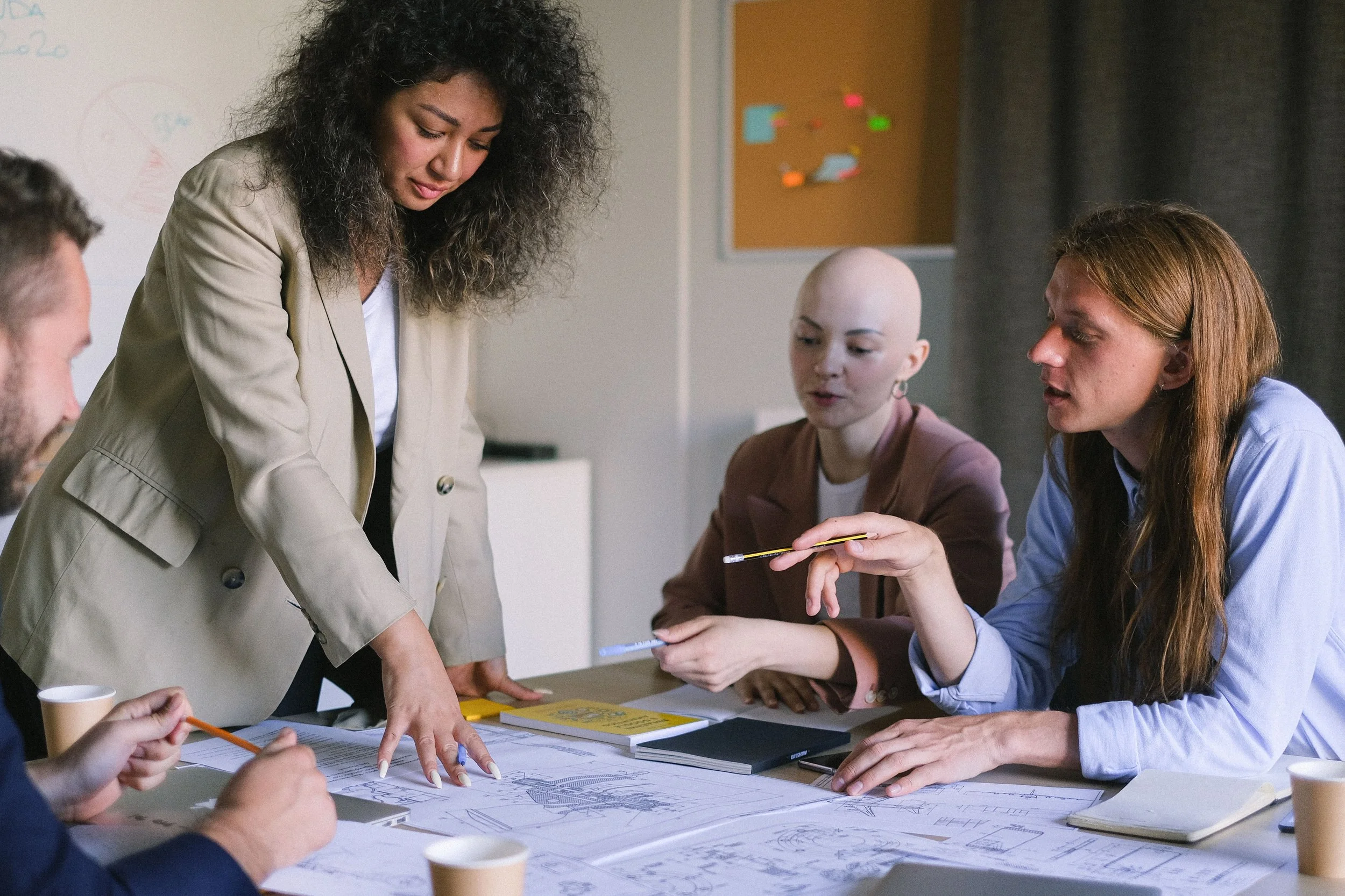 A group of five people working together at a table with architectural plans, notebooks, and pens. One woman with curly hair is standing and pointing at the plans while others observe and discuss.