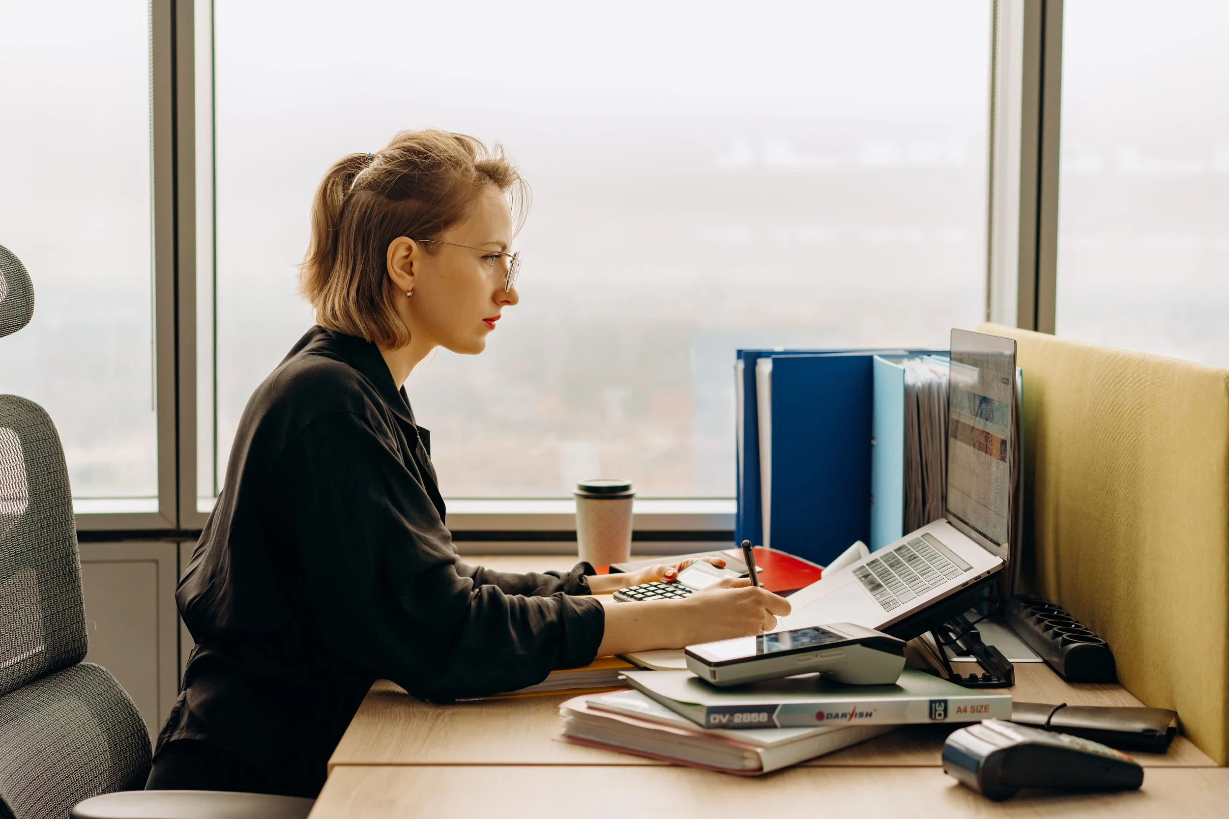 A woman with glasses working at a desk in front of a computer, surrounded by office supplies, with large windows in the background.