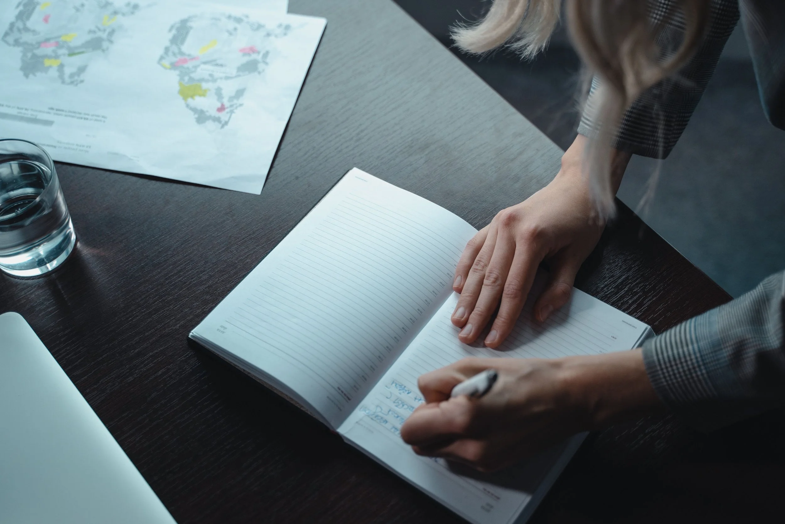 Person writing in a lined journal at a dark wooden desk, with a glass of water, a world map, and a closed silver laptop nearby.