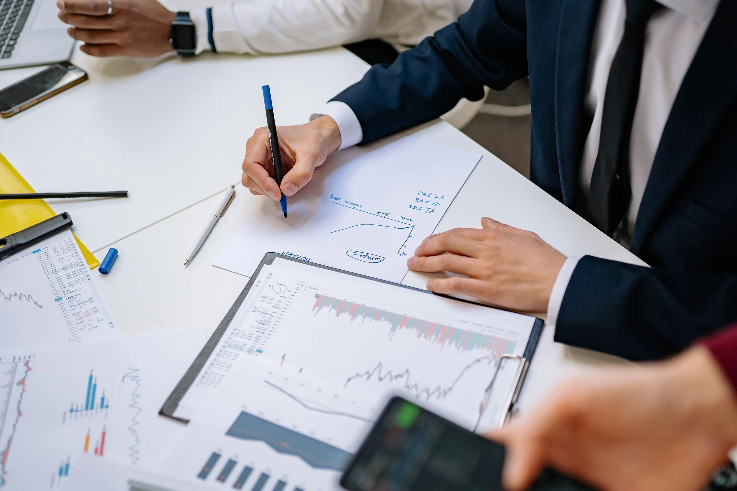 People sitting at a white table working on financial charts and graphs, using pens, laptops, and smartphones.