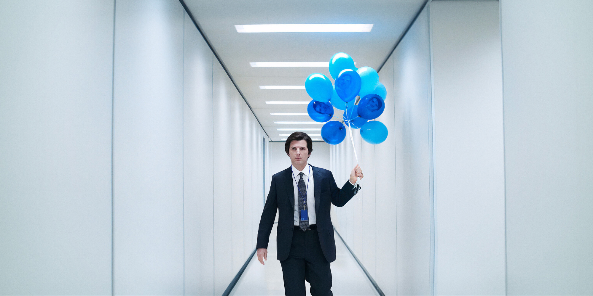 A man in a suit, Mark S. from Severance, holding blue balloons in a sterile white office, representing the performative and "perfect" appearance of the fawn response.