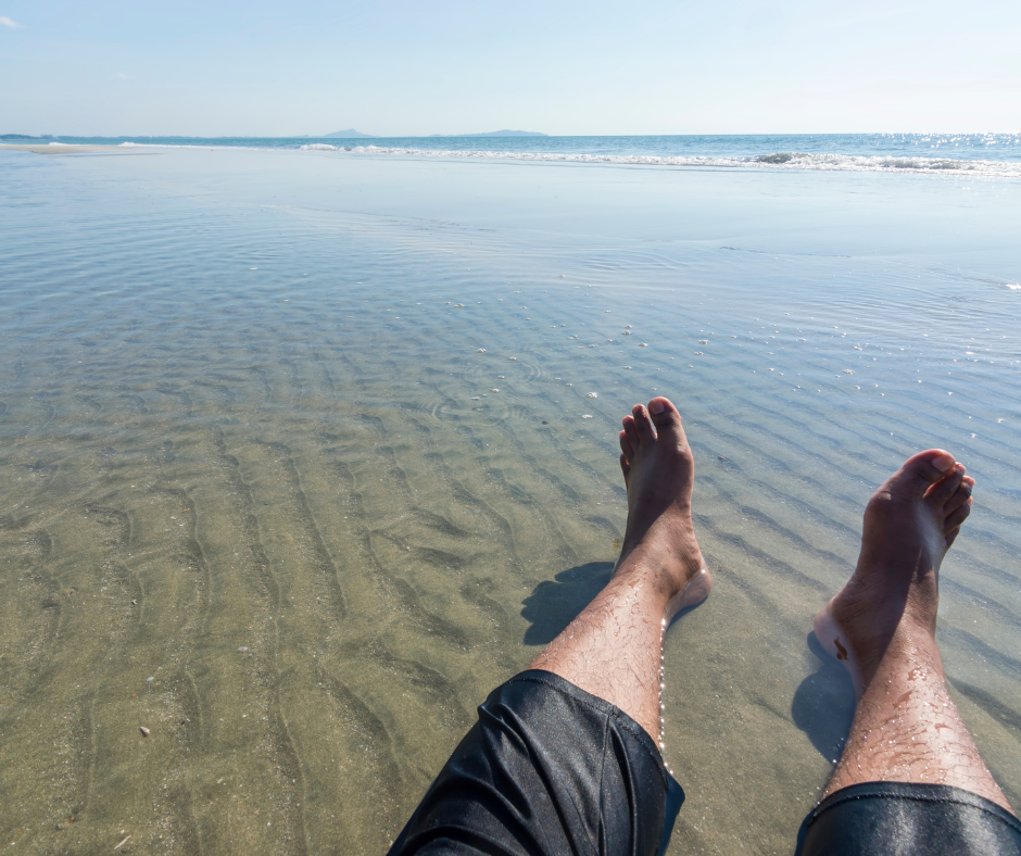 A person sitting with their feet in shallow ocean water on a beach, representing the letting go of effort in ACT for insomnia.