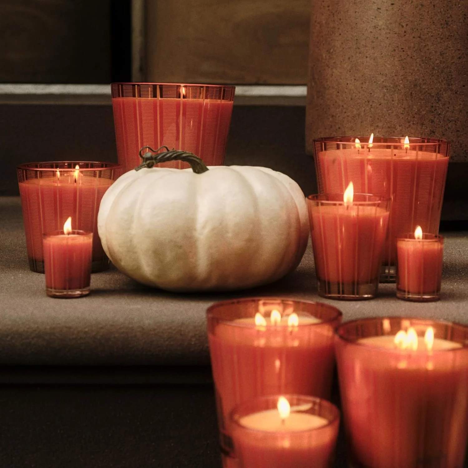 A white pumpkin surrounded by lit orange candles in glass holders on a dark surface, with a background of a brown textured wall and a partial pillar.