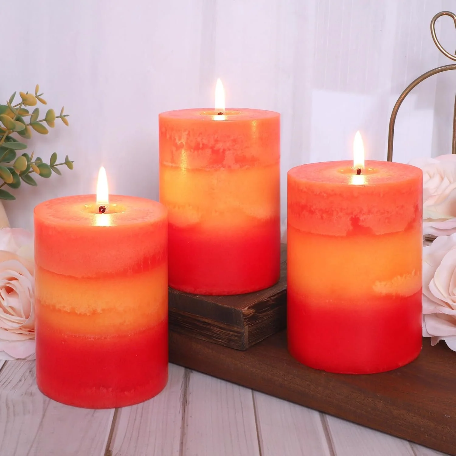 Three lit orange candles on a wooden board surrounded by white roses and greenery against a white background.