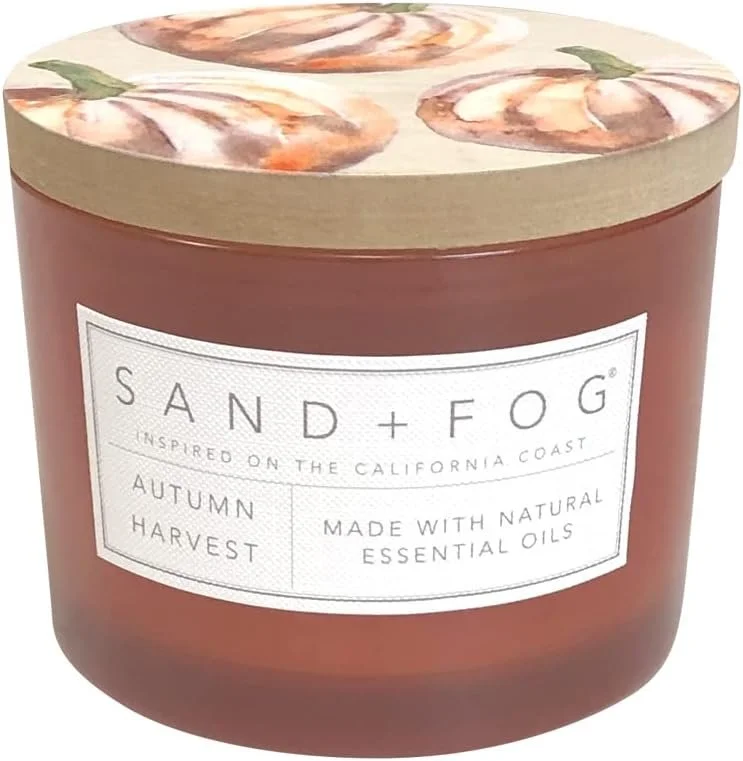 A candle in a brown jar labeled 'Sand + Fog' with scent 'Autumn Harvest,' made with natural essential oils, inspired by the California coast. The lid has a floral design with peach and green colors.