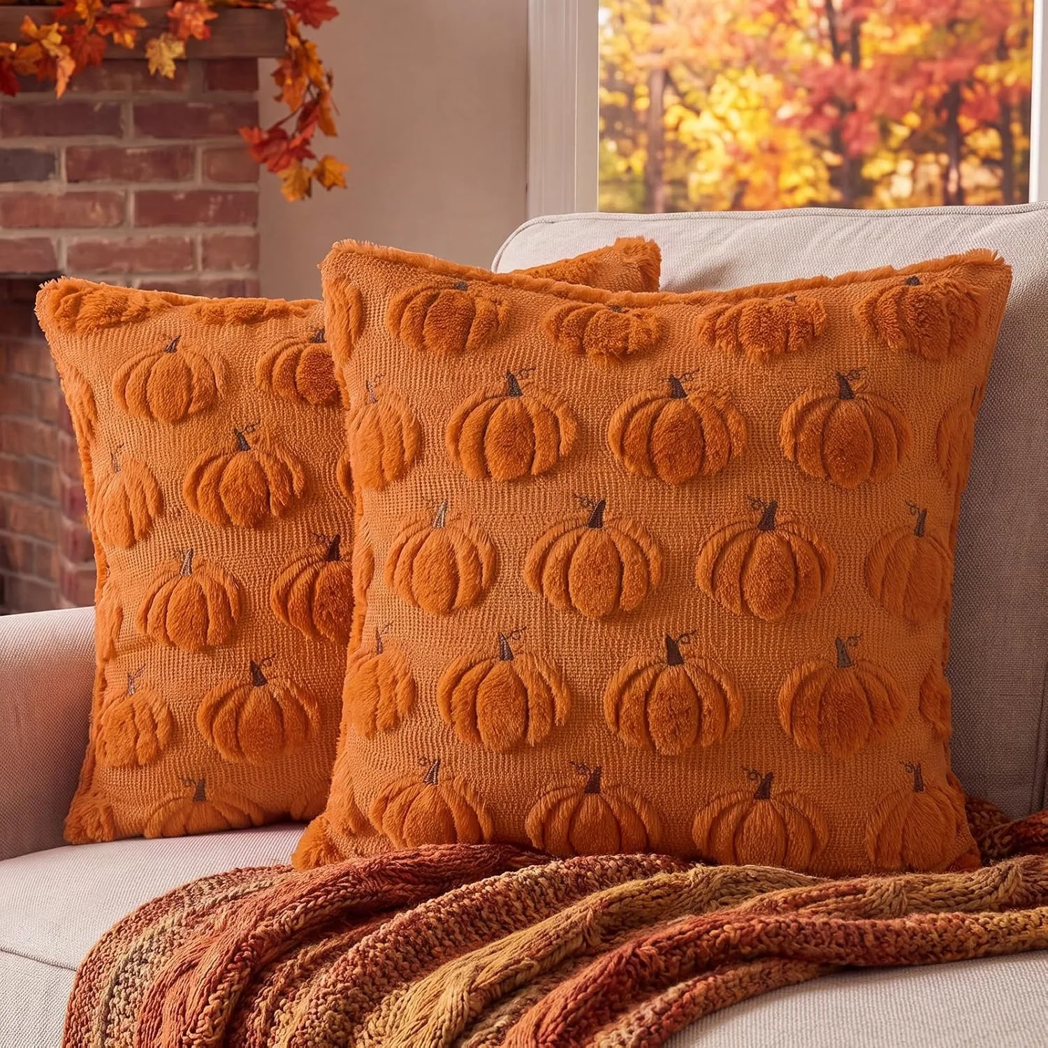 Two orange throw pillows with textured pumpkin embroidery sit on a light-colored sofa. A cozy, multicolored blanket is draped over the sofa in front of the pillows. In the background, there are autumn leaves outside the window, and a brick fireplace decorated with a garland of fall-colored leaves.