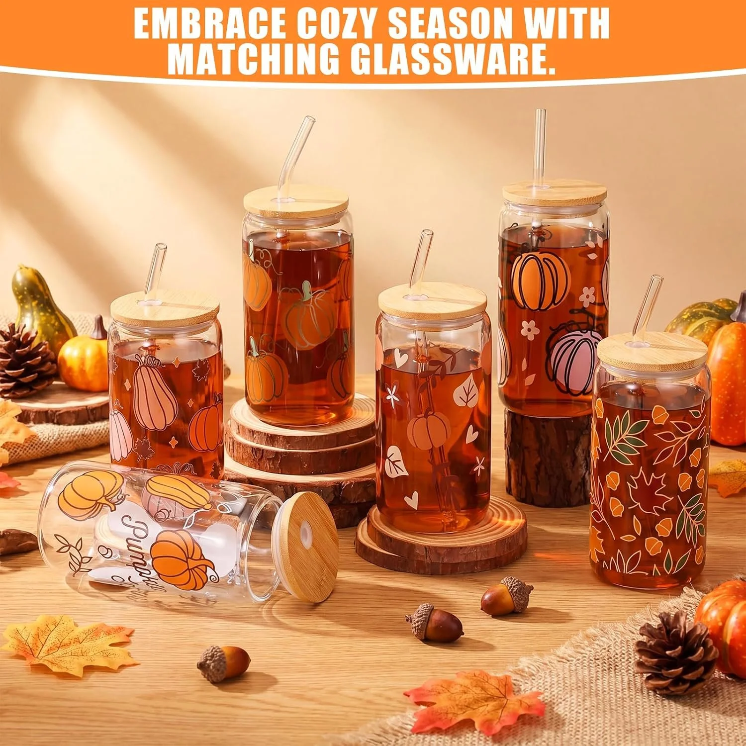 Five glass jars with autumn-themed pumpkin designs, filled with an amber-colored beverage, are displayed on a wooden surface with autumn leaves and pine cones; one jar is lying on its side with a wooden lid beside it. Additional pumpkins and greenery are in the background. The text reads 'Embrace cozy season with matching glassware'.