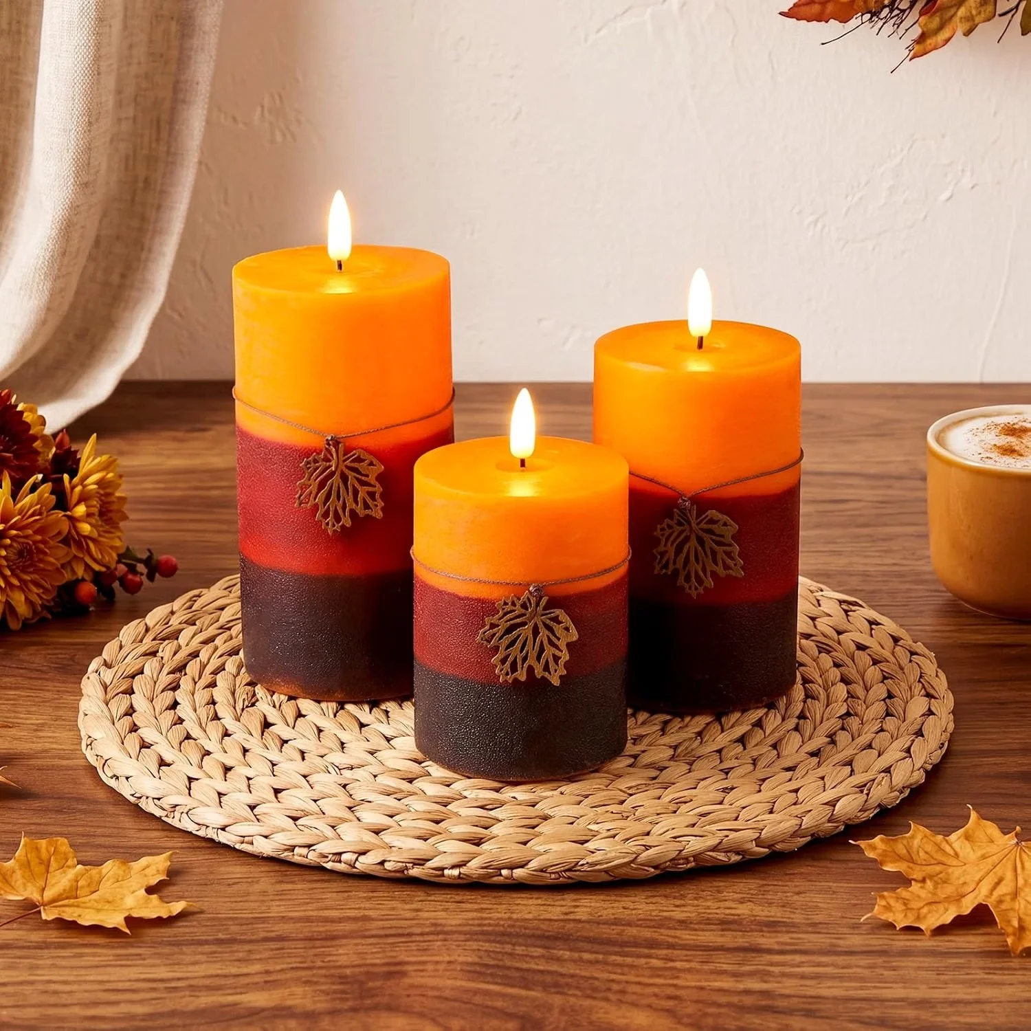 Three orange and black gradient candles with leaf decorations, arranged on a woven mat on a wooden table with autumn leaves and a cup of coffee in the background.