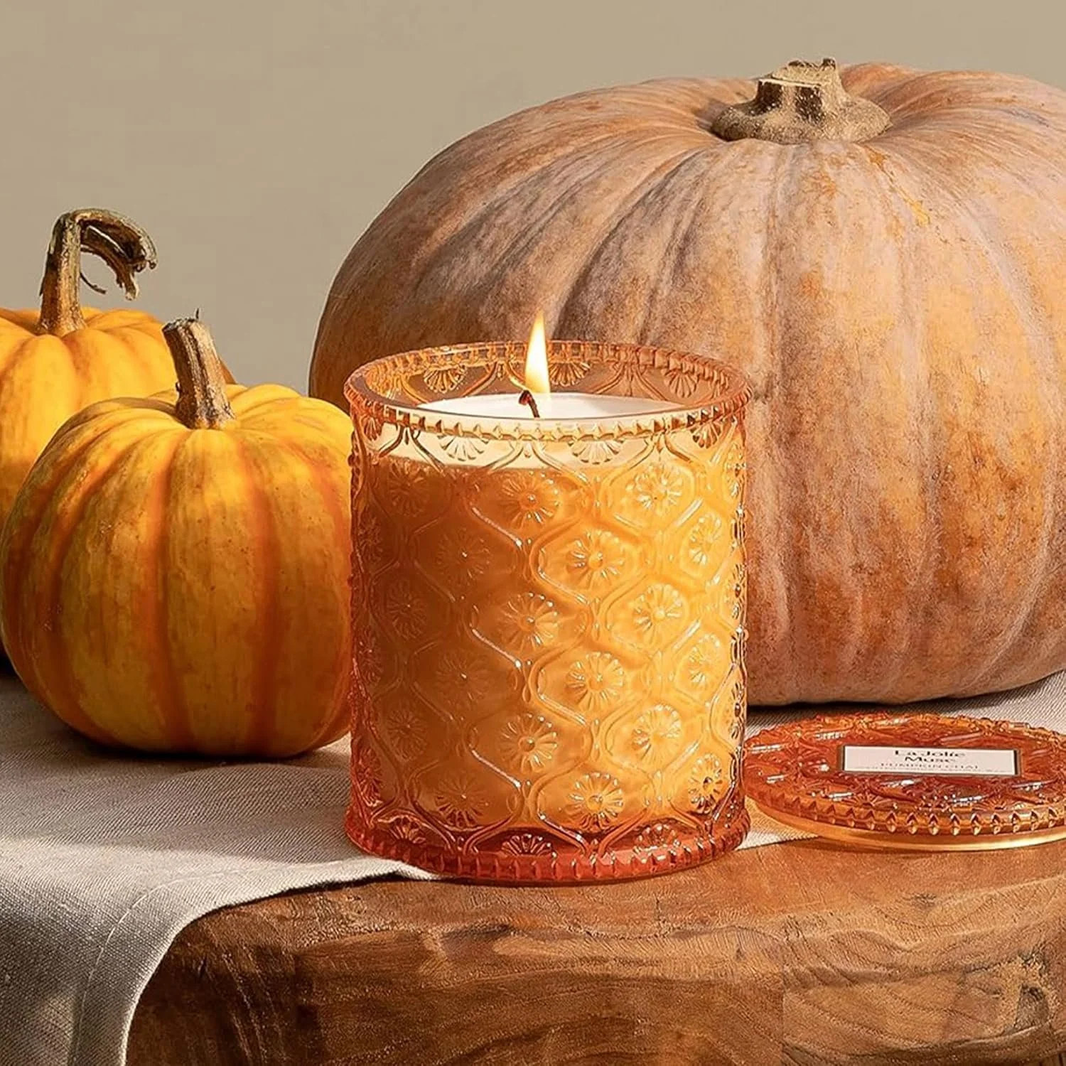 A lit orange candle in an intricate glass holder surrounded by pumpkins, including two small orange pumpkins and a larger white pumpkin on a wooden surface with a beige background.