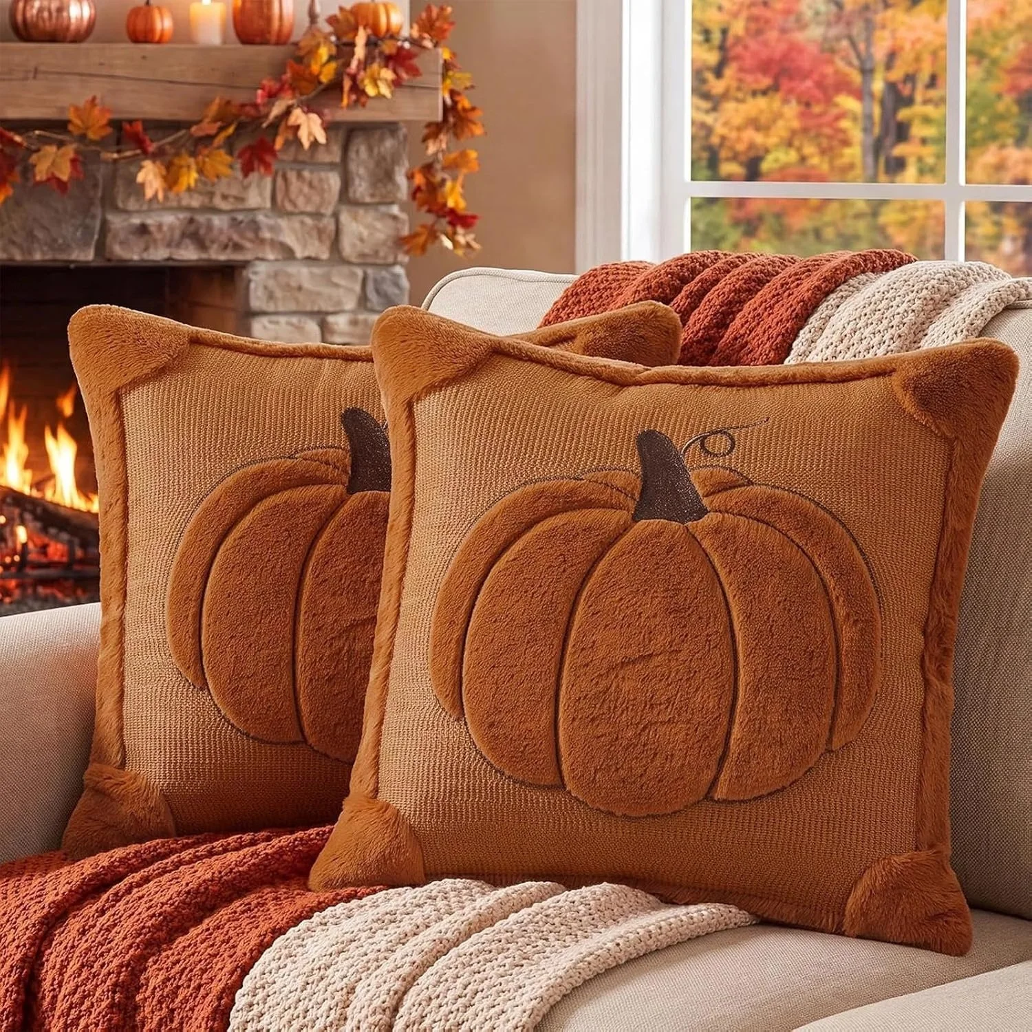 Two decorative pillows with pumpkin designs on a beige couch, autumn-themed blankets, a fireplace with a wooden mantel, and fall-colored trees visible through a window.