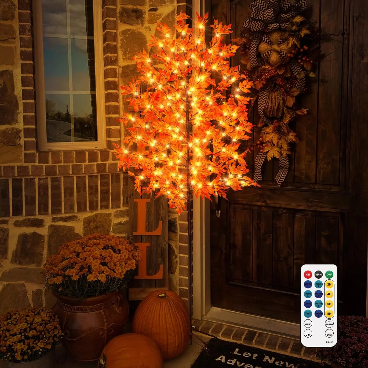 An illuminated artificial orange maple tree with lights, situated in front of a brick wall and a wooden door. Decorated autumnal accessories include a goblet, pumpkins, and potted chrysanthemums. A large orange letter "L" and a small pumpkin are visible on the ground, and a welcome mat with partial text can be seen at the bottom of the image.
