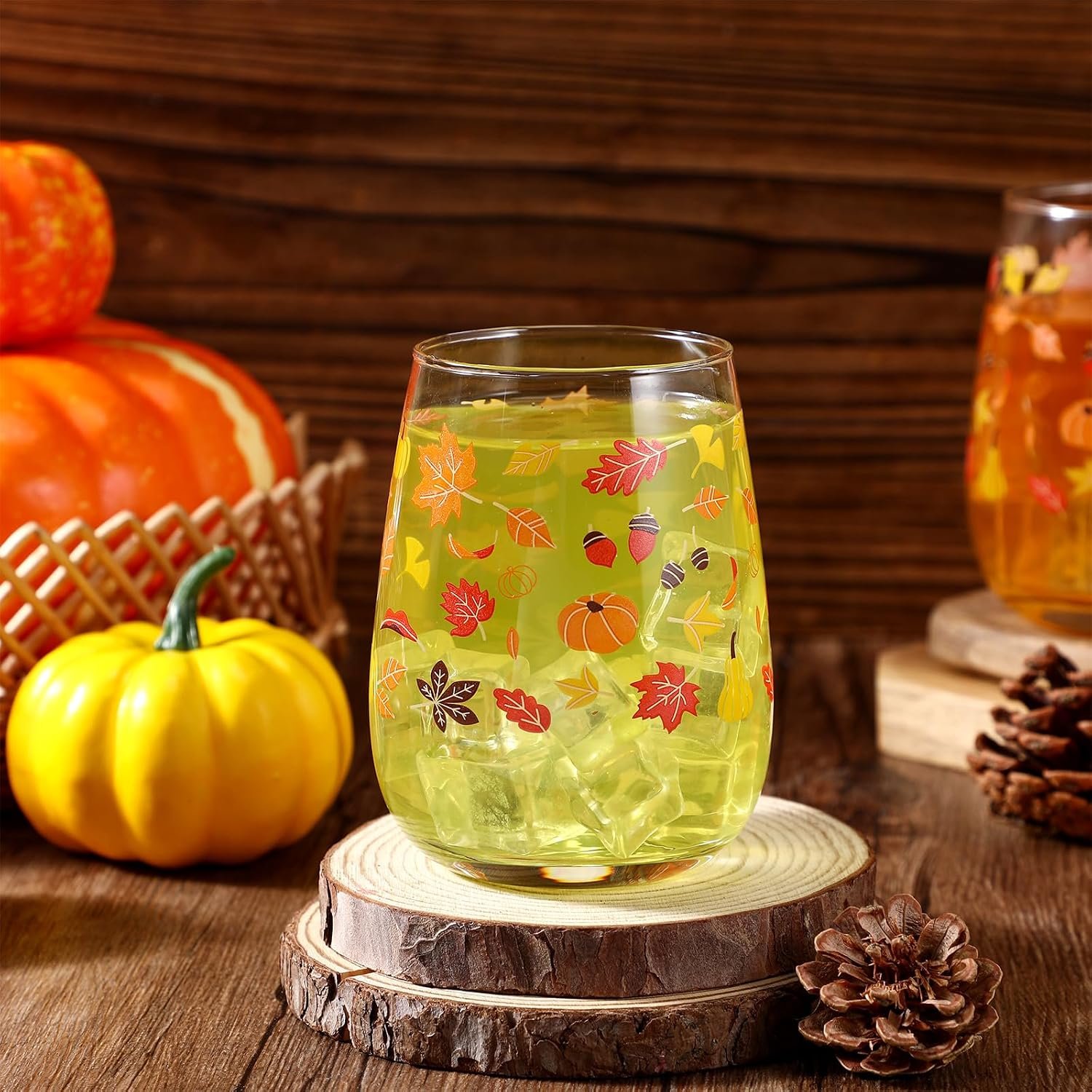 A glass of yellow-green beverage with ice and autumn leaf decorations, placed on a wooden slab, surrounded by pumpkins and pinecones, with a wooden background.