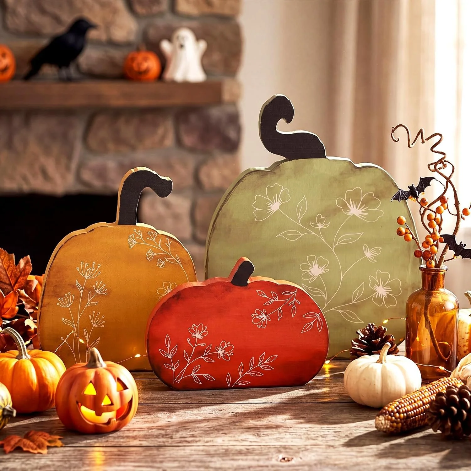 A Halloween decoration display with painted pumpkins, a Jack-o'-lantern, fall leaves, pinecones, and a vase with decorative branches and bats, set on a wooden surface in front of a cozy fireplace with holiday figurines.