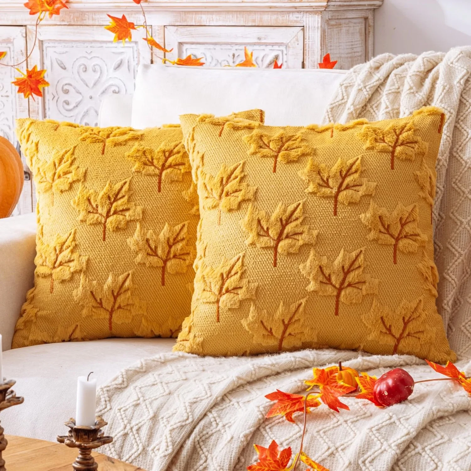 Decorative autumn-themed pillows on a cream-colored sofa, with scattered orange and red fall leaves and a red apple on a cream blanket.