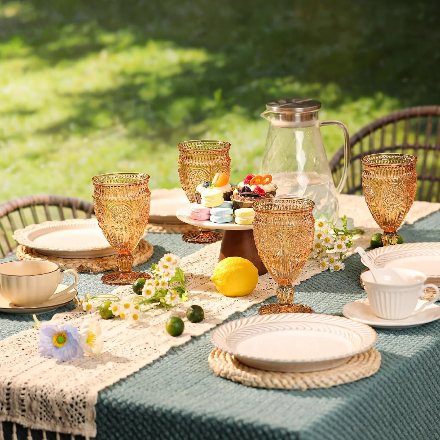 A table set for an outdoor meal with pink vintage glasses, white and pastel dishes, a carafe, colorful macarons, fresh berries, daisies, and a lemon, on a blue and beige tablecloth.
