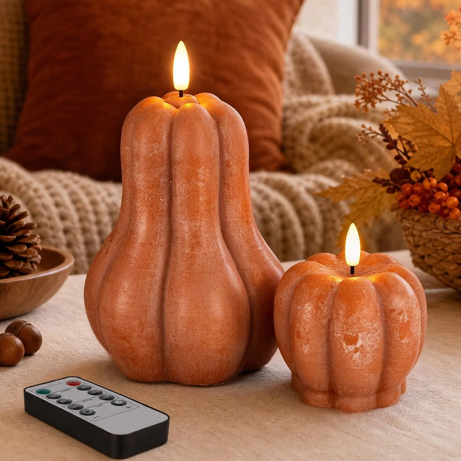 Two pumpkin-shaped candles with electric flames on a table, surrounded by autumn decor including pinecones, berries, and leaves.