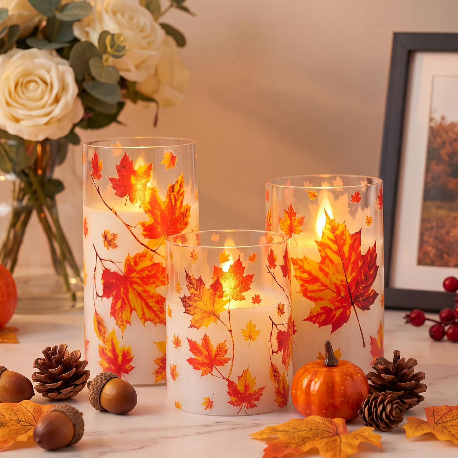 Decorative fall-themed candles with autumn leaves design, surrounded by small pumpkins, pinecones, acorns, and fall leaves, with a flower arrangement and framed picture in the background.