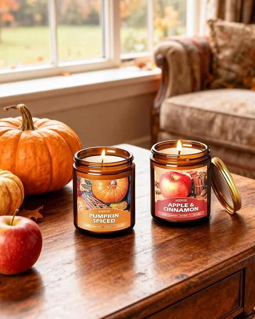 Two lit candles in amber jars on a wooden table next to pumpkins and apples, with a sofa and window in the background.