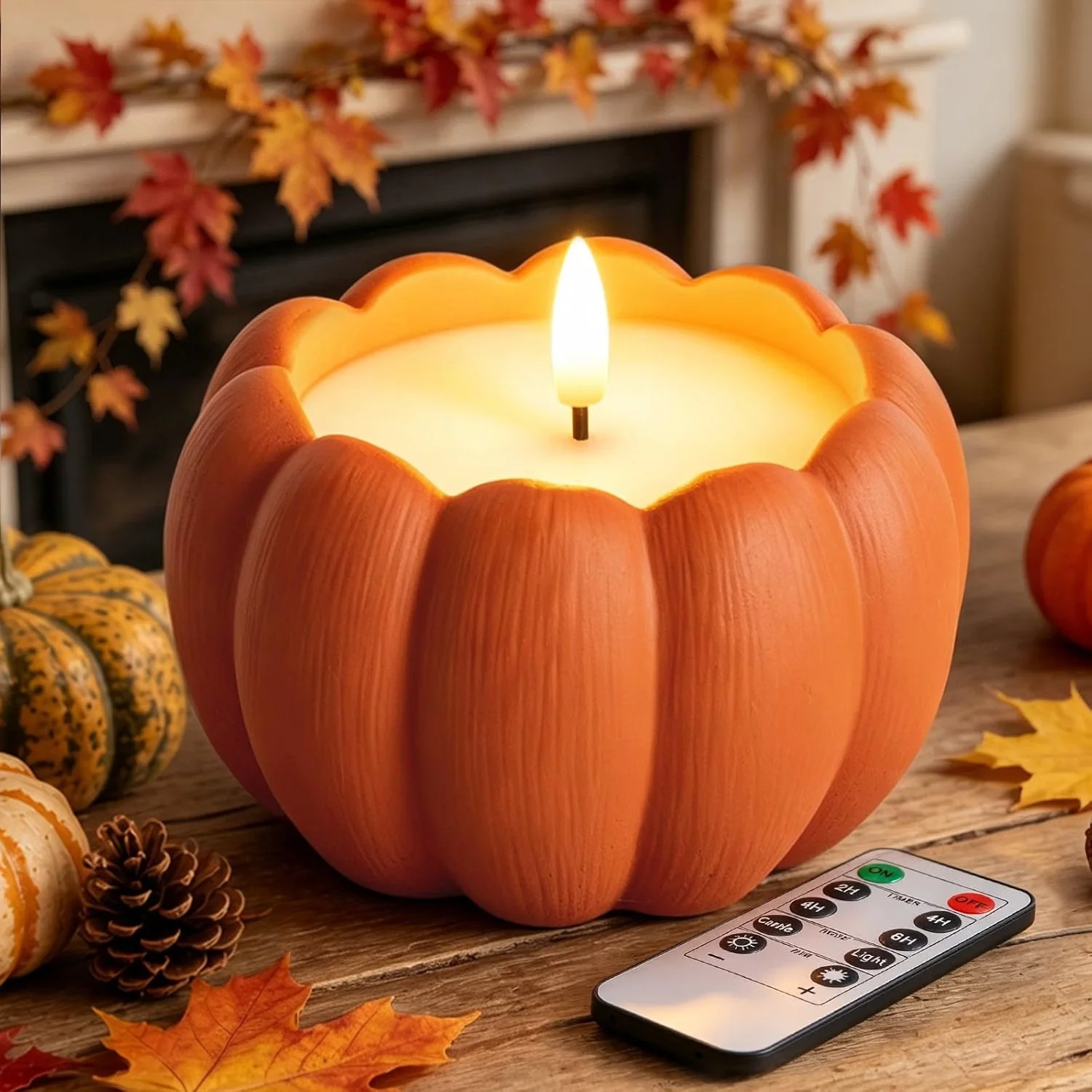 A pumpkin-shaped candle with a lit flame on a wooden table, surrounded by autumn leaves, small pumpkins, pine cones, and a remote control.
