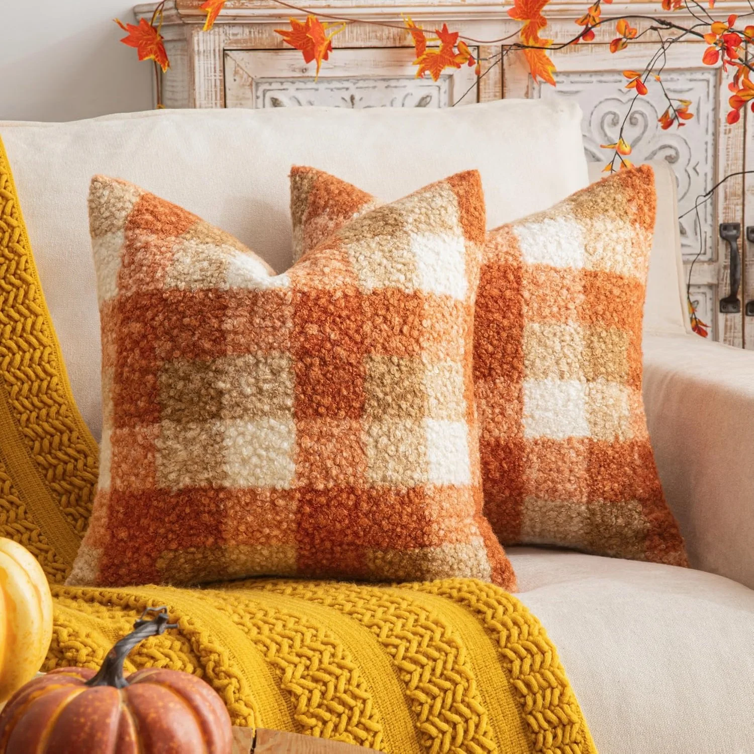 Decorative fall-themed interior with two orange patterned pillows on a white sofa, a yellow knit throw, and small pumpkins, with orange and yellow autumn leaves on the wall.