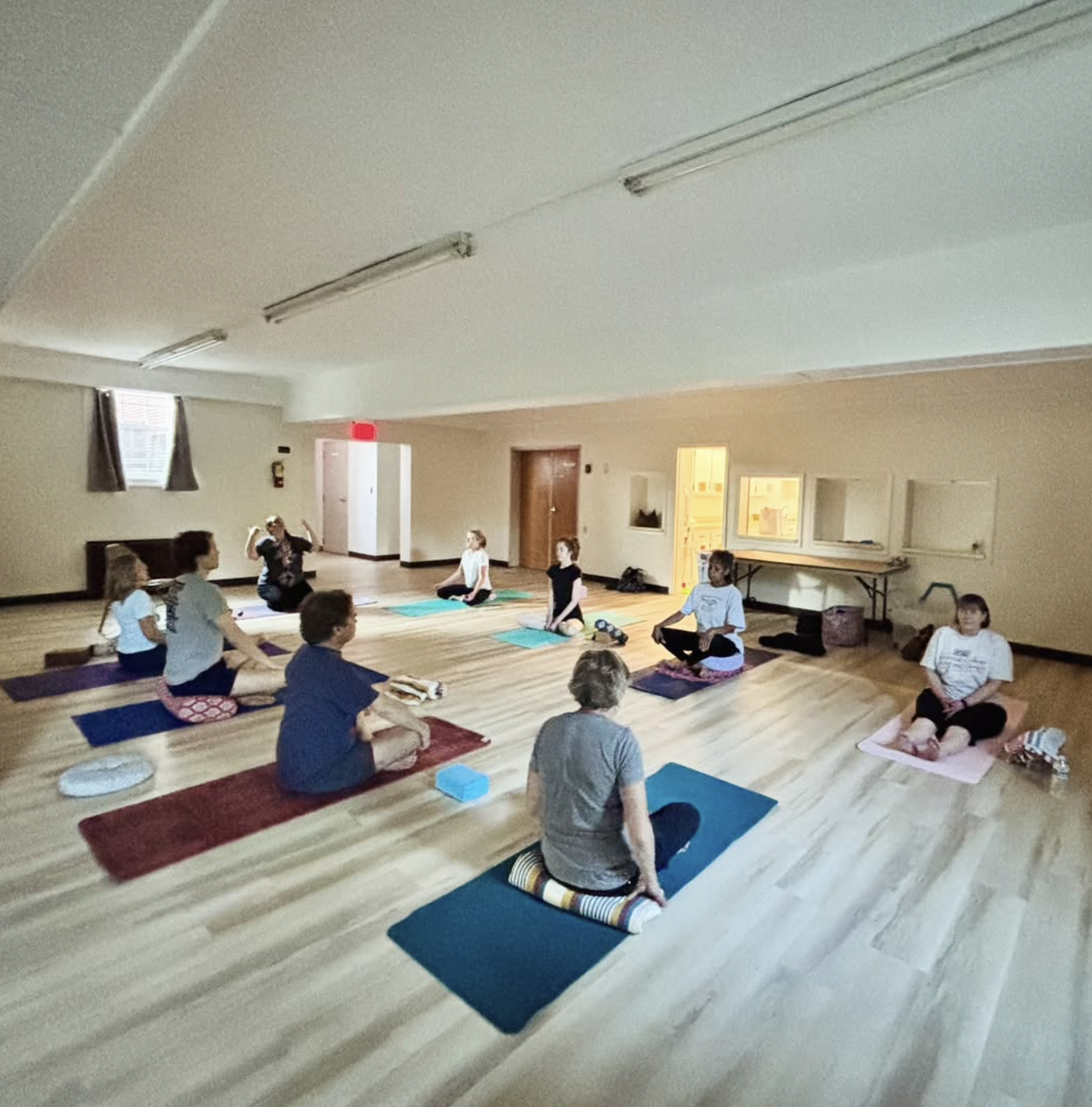 Group of people participating in a yoga class in a spacious room with wooden floor and high ceiling, sitting on yoga mats in a cross-legged position, some with eyes closed, instructor leading the session.