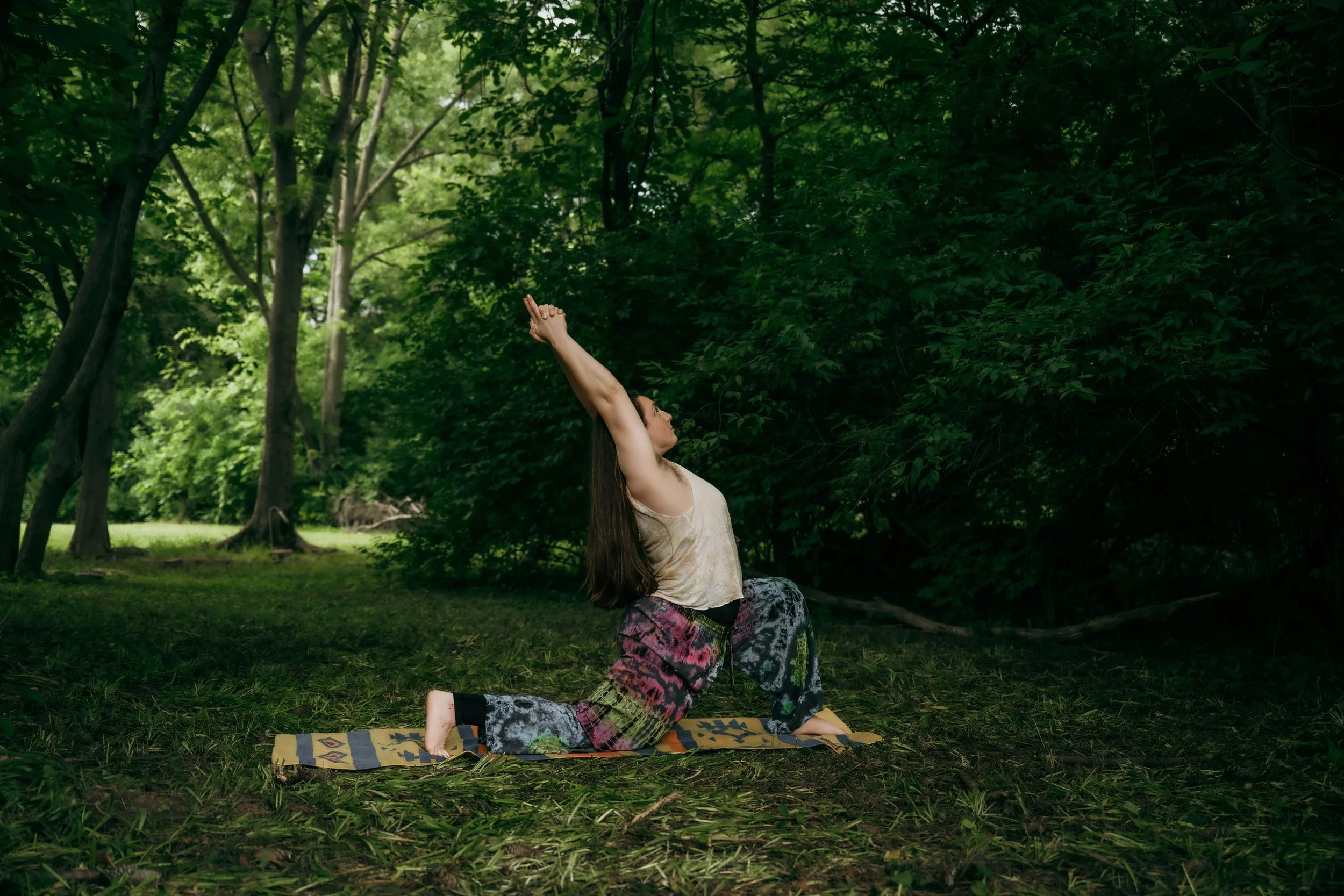 A woman practicing yoga outdoors in a wooded area, performing a child pose with her arms extended in the air on a yoga mat.