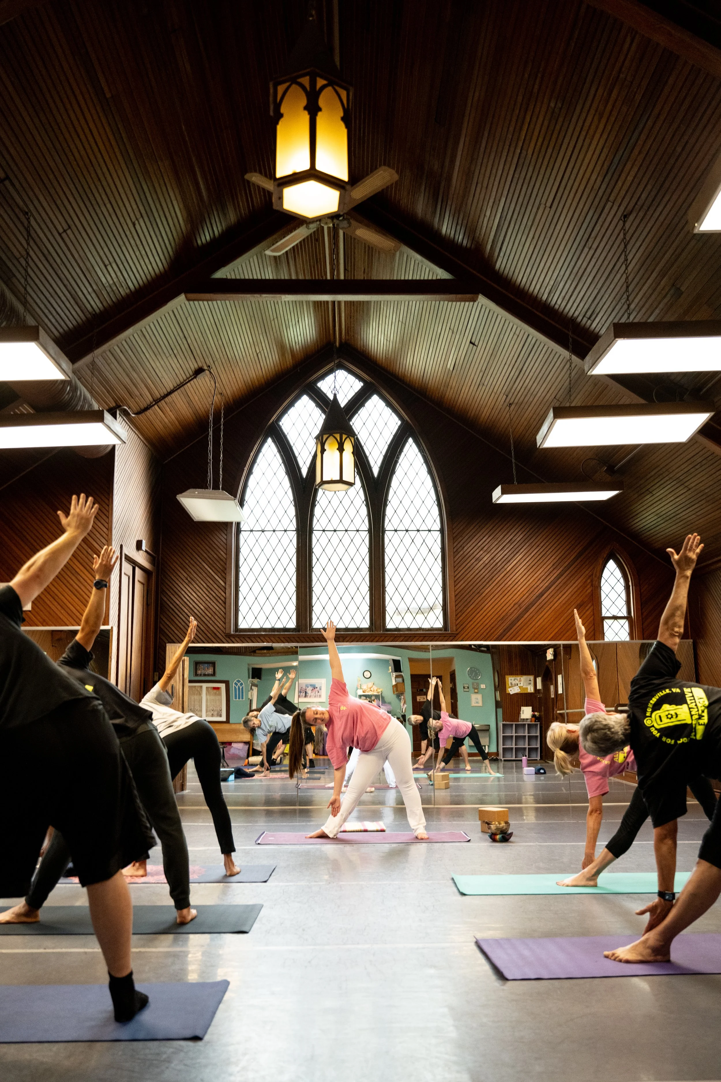 Yoga class in a large, wooden-ceiling room with stained glass windows, participants on mats doing stretches, and a trainer assisting in the front.