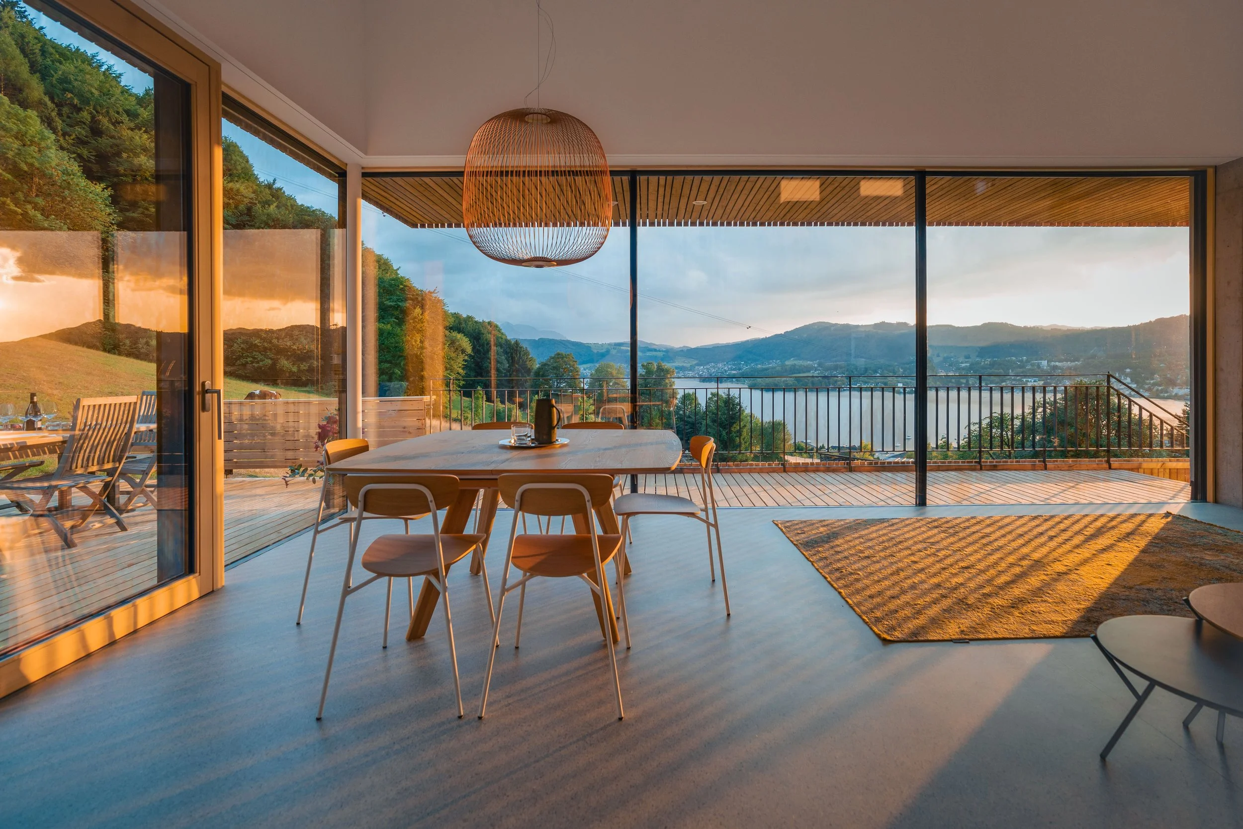 Innenraum eines Hauses mit großer Fensterfront, Blick auf Berge und Wasser, Essbereich mit sechs Stühlen um einen runden Tisch, im Raum ein Holzzuhhängelicht und Teppich auf hellem Boden.