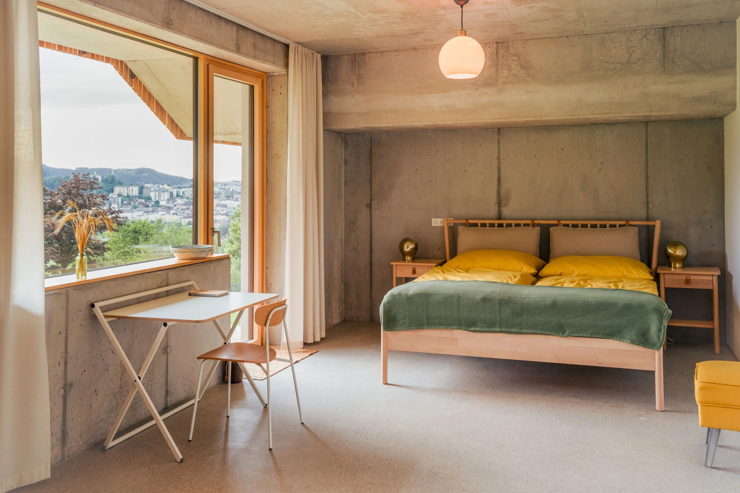 Schlafzimmer mit einem Bett, zwei Nachttischen, einem Schreibtisch am Fenster, Blick auf eine Stadt und Berge im Hintergrund, Modernes Dekor, natürliche Farben.