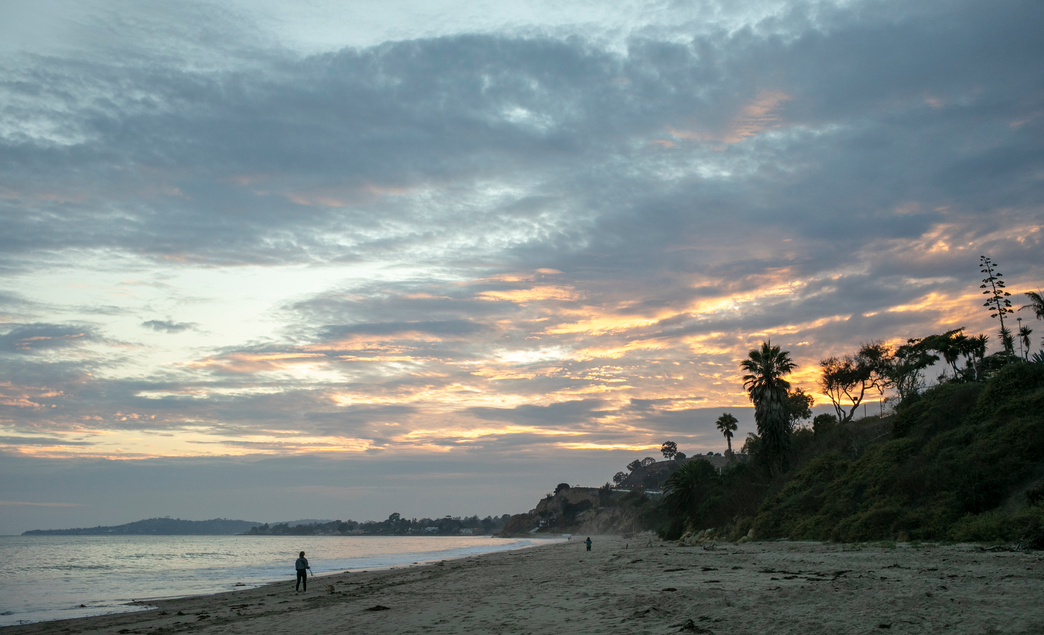 Sunset over a beach with a few trees and people walking along the shoreline.