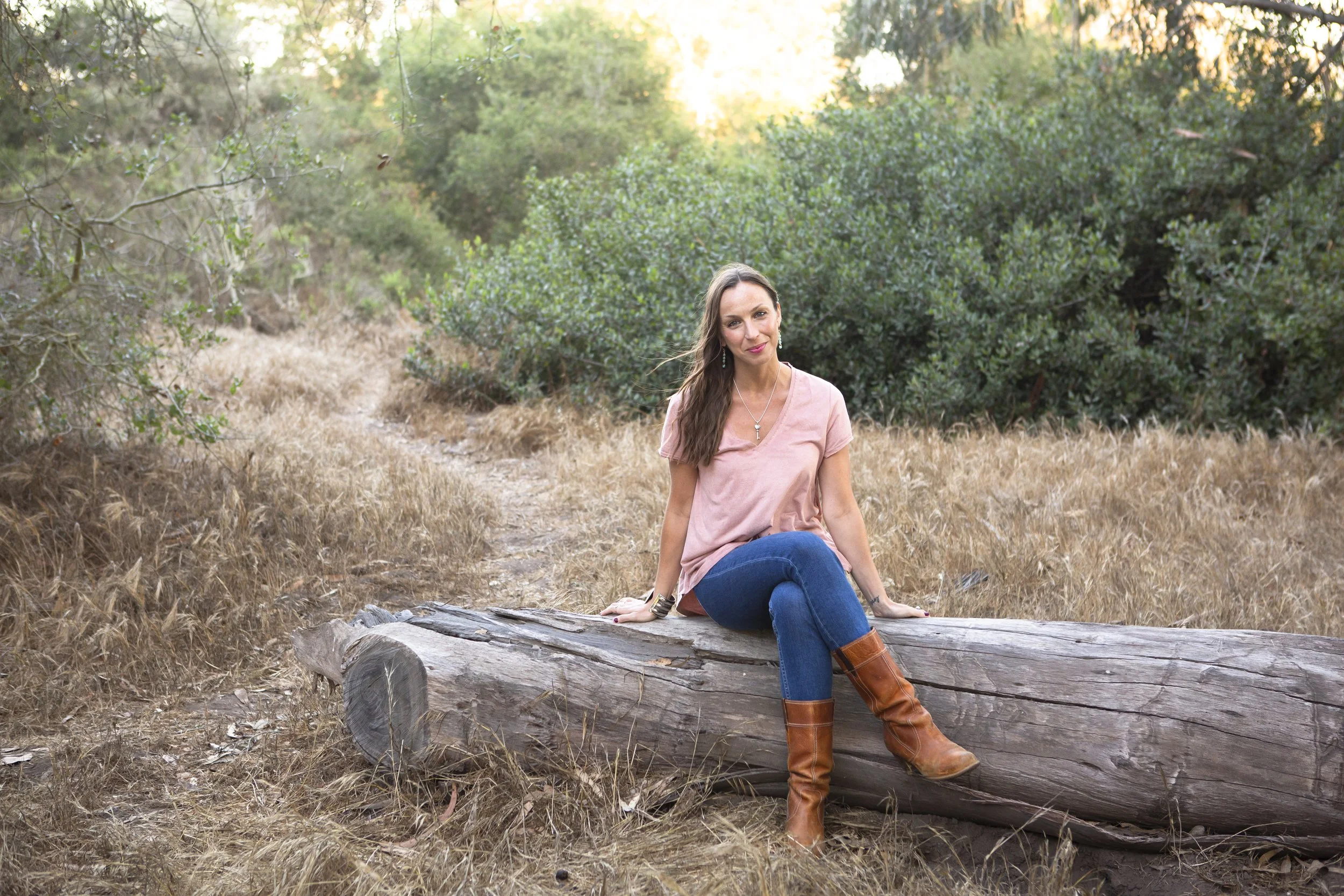 A woman sitting on a fallen log in a natural outdoor setting with dry grass and green bushes, during sunset.