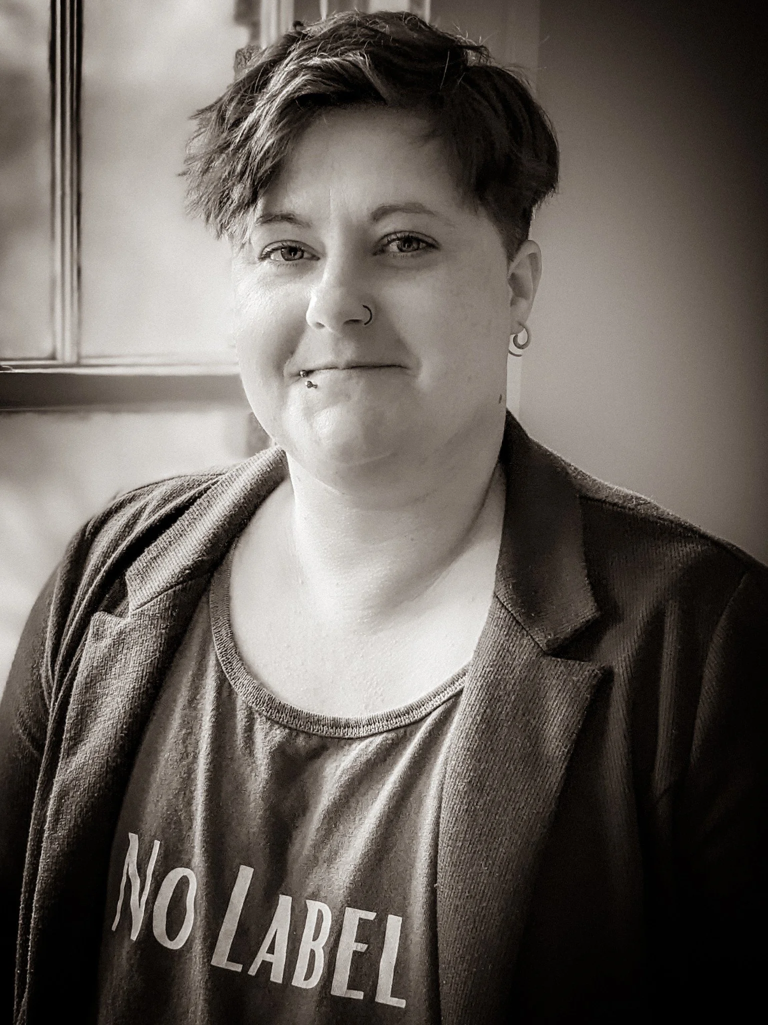 A person with short, tousled hair and light-colored eyes, wearing earrings, a nose ring, and a labret piercing, smiling slightly, dressed in a T-shirt with 'NO LABEL' text and a dark jacket, in a room with a wooden cabinet.
