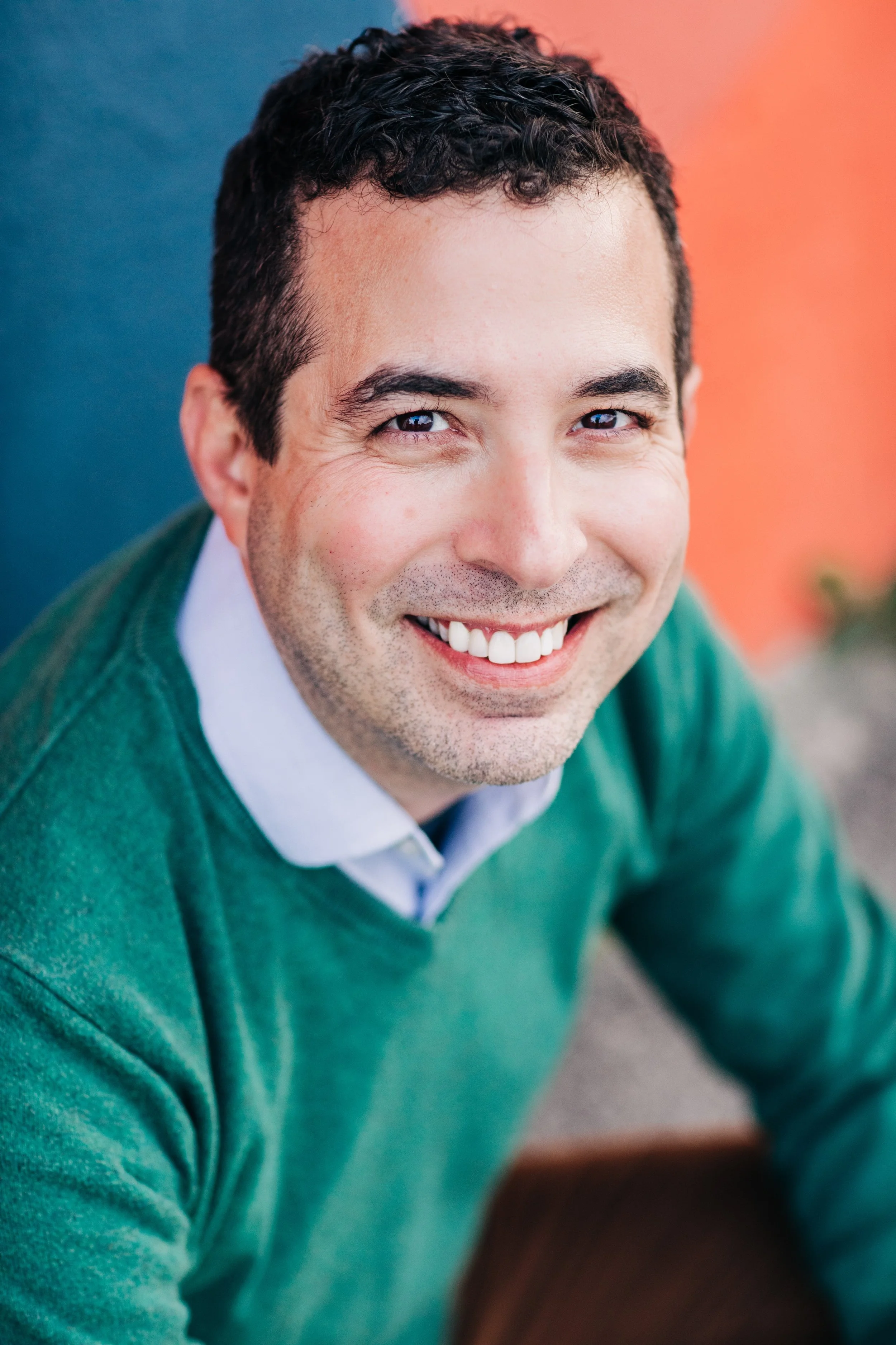 Close-up of smiling man in green sweater and white shirt against colorful background.