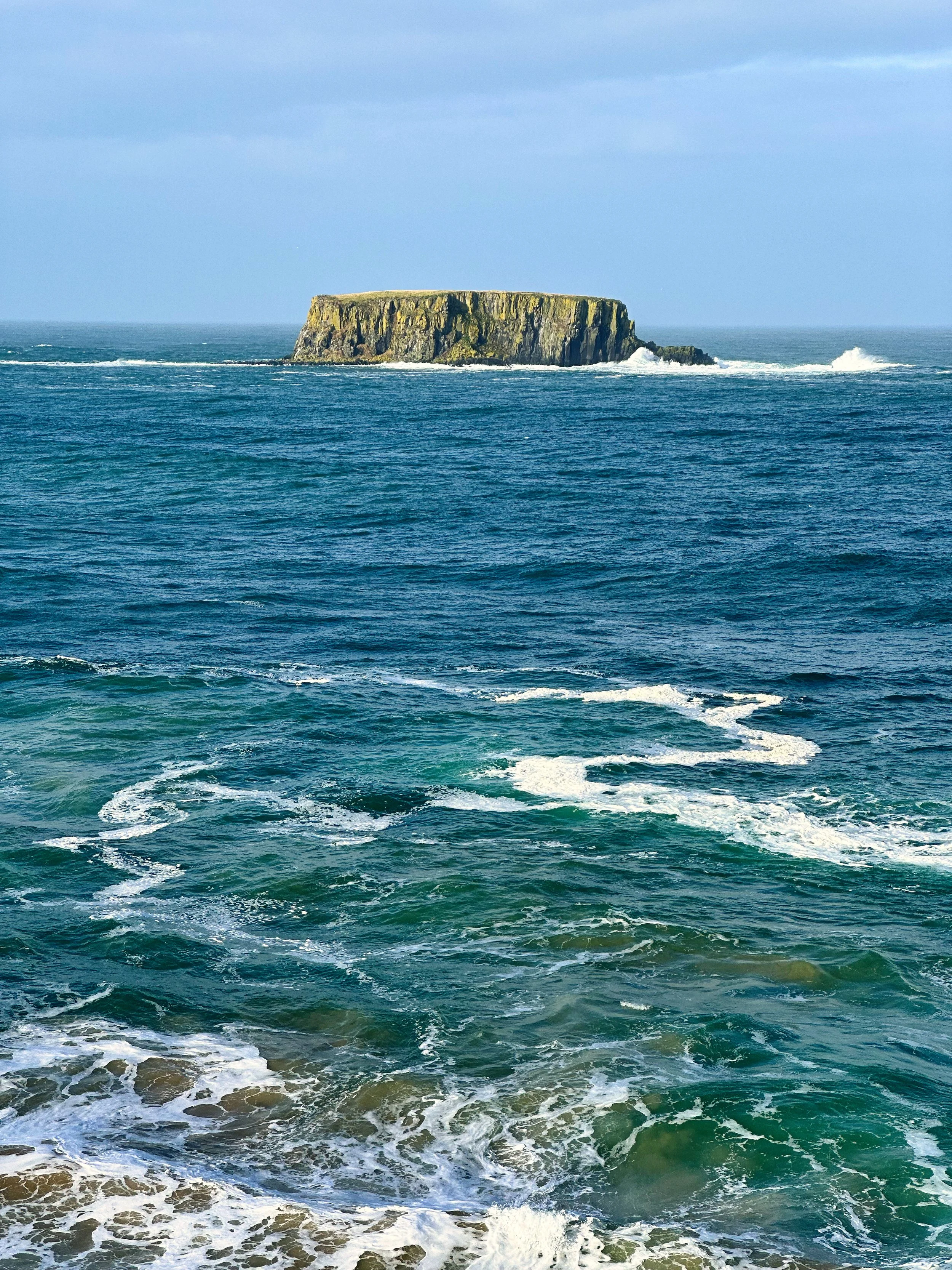 A large, flat-topped island or rock formation in the ocean, with waves in the foreground and a cloudy sky above.