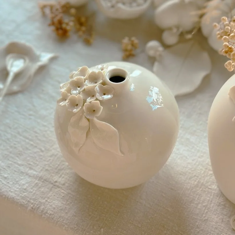 Vase blanc en céramique avec décoration florale en relief, placé sur une surface en tissu. Autres objets similaires et fleurs en arrière-plan.