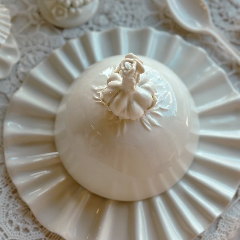 Un dessert rond blanc avec un décor en forme de chapeau de cupcake, placé sur une assiette blanche avec un motif en relief. Autour, une nappe en dentelle et une petite cruche en porcelaine.