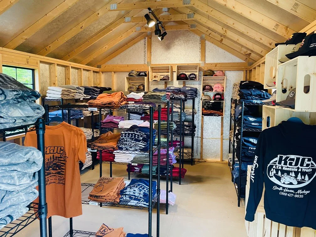 Interior of KJC's gift shop with shelves and racks filled with T-shirts, hats, and hoodies in a rustic wooden space.