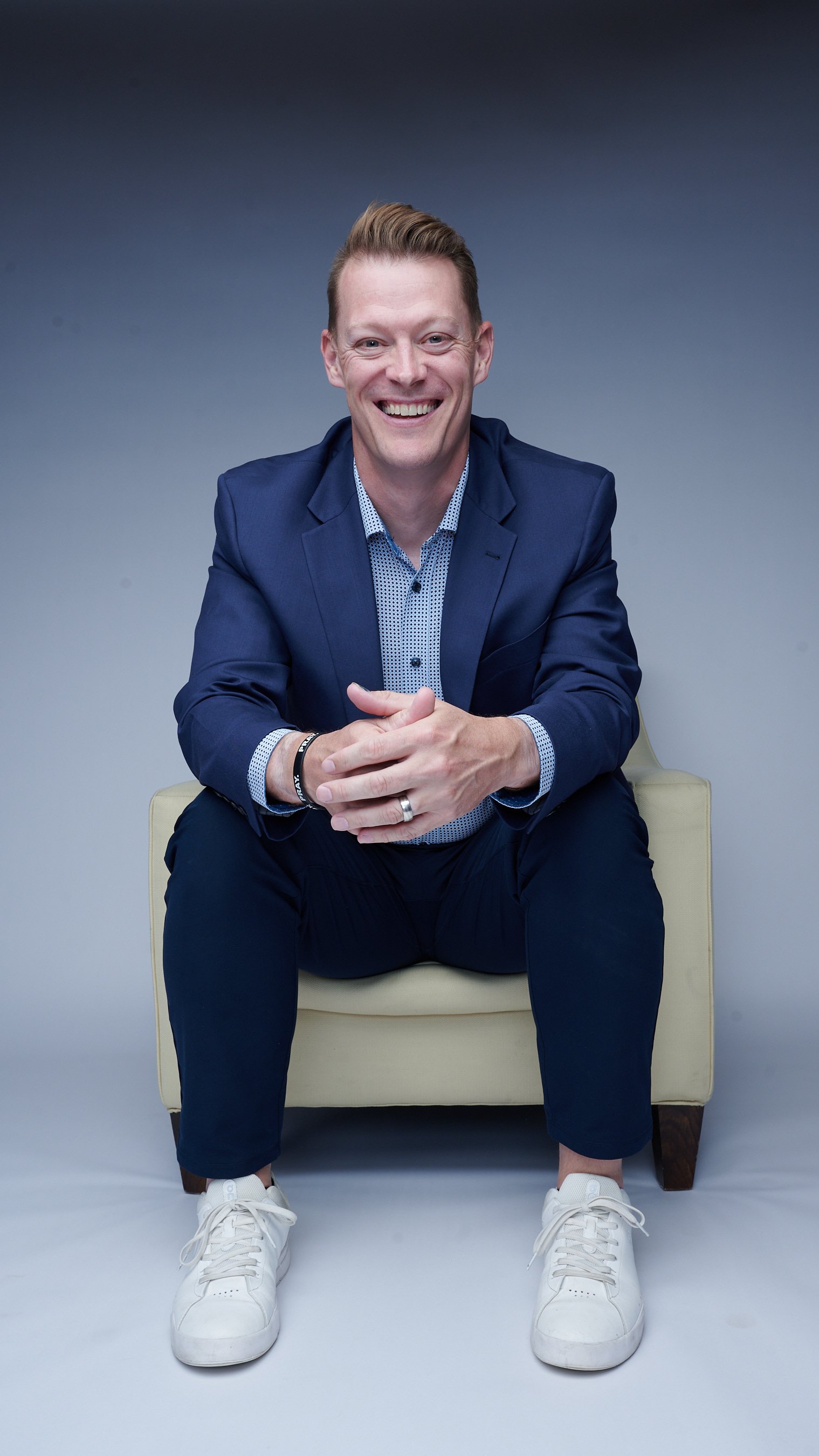 A man with short blonde hair sitting on a white armchair against a gradient gray background, wearing a navy blazer, a patterned shirt, dark trousers, and white sneakers, smiling at the camera with hands clasped.