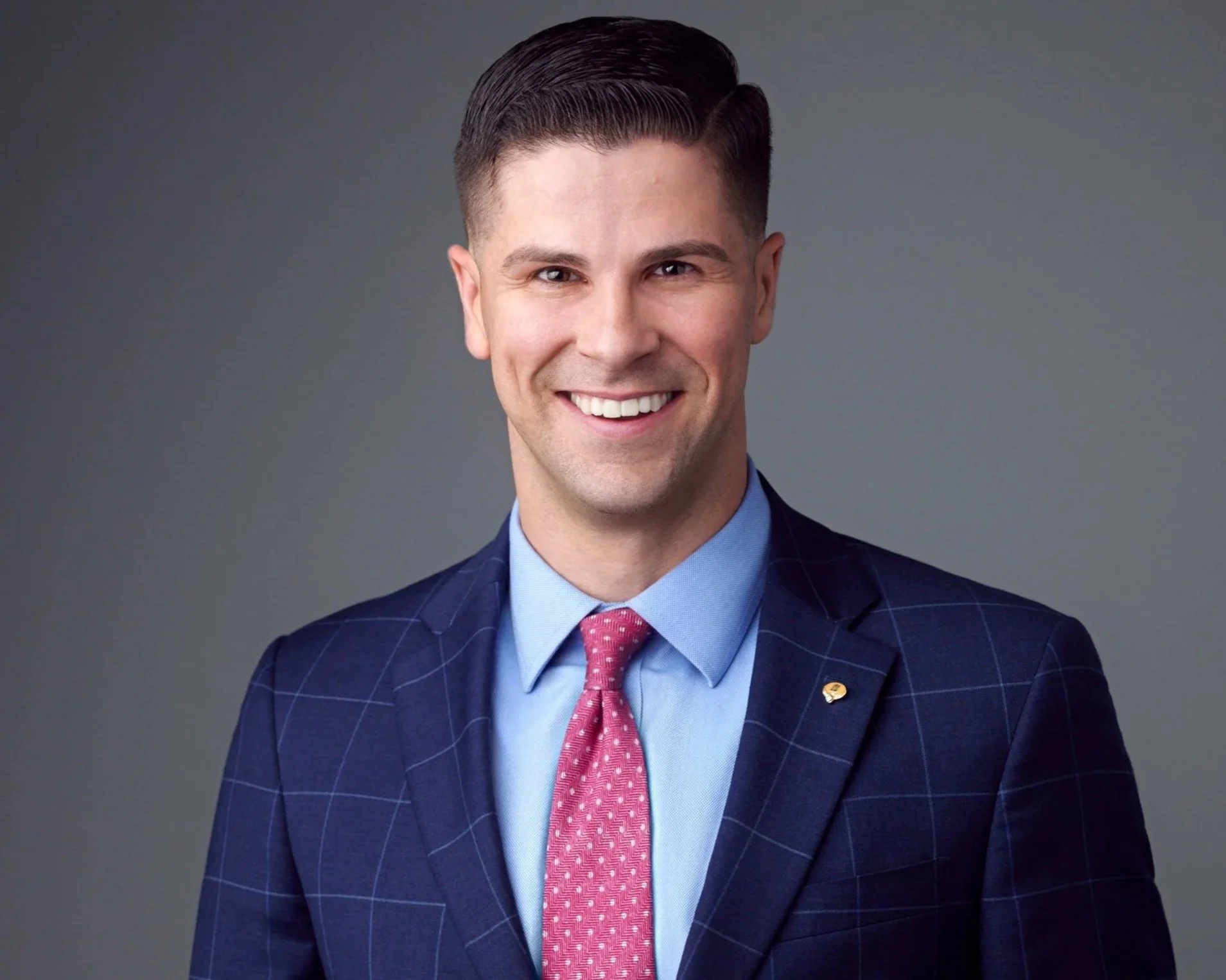 A man in a dark blue checked suit, light blue shirt, and pink tie, smiling at the camera against a gray background.