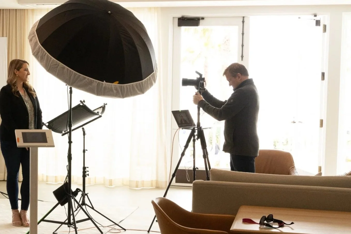 A woman being filmed in a bright indoor setting with professional lighting and filming equipment, including a large umbrella-style softbox, a camera on a tripod, and a laptop.