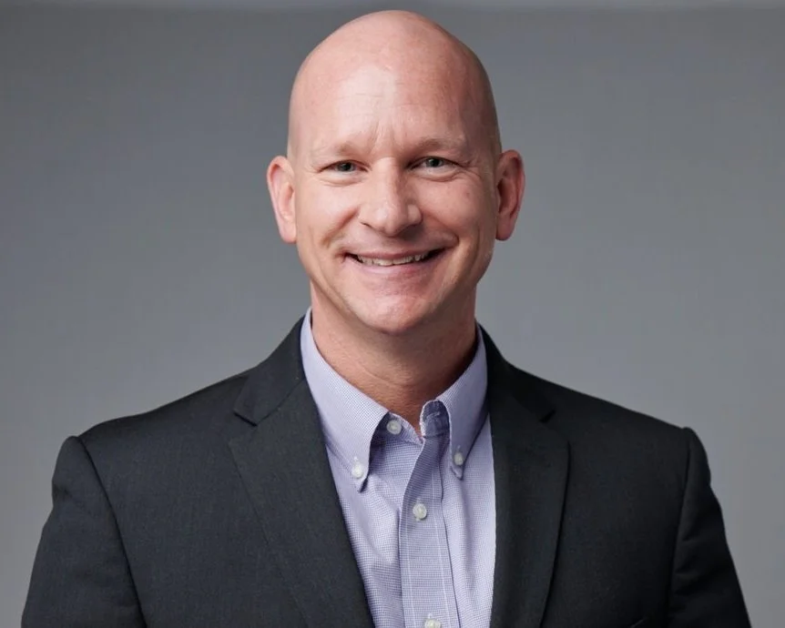 A man in a dark suit and light purple collared shirt smiling in front of a plain gray background.