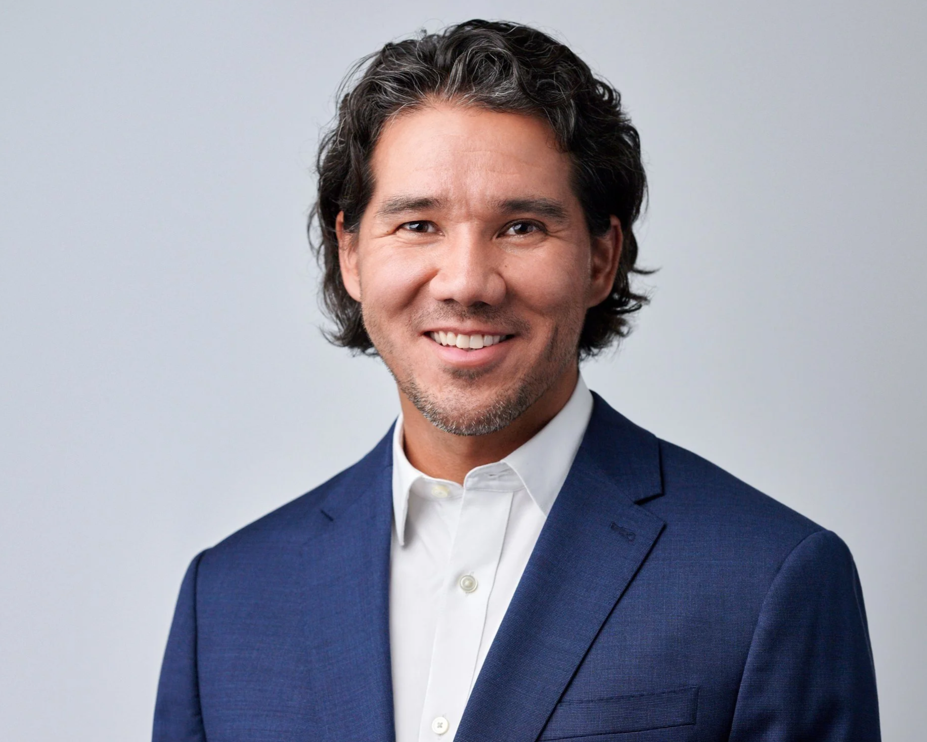 Headshot of a man with wavy dark hair, smiling, wearing a navy blue suit and white shirt, against a light grey background.