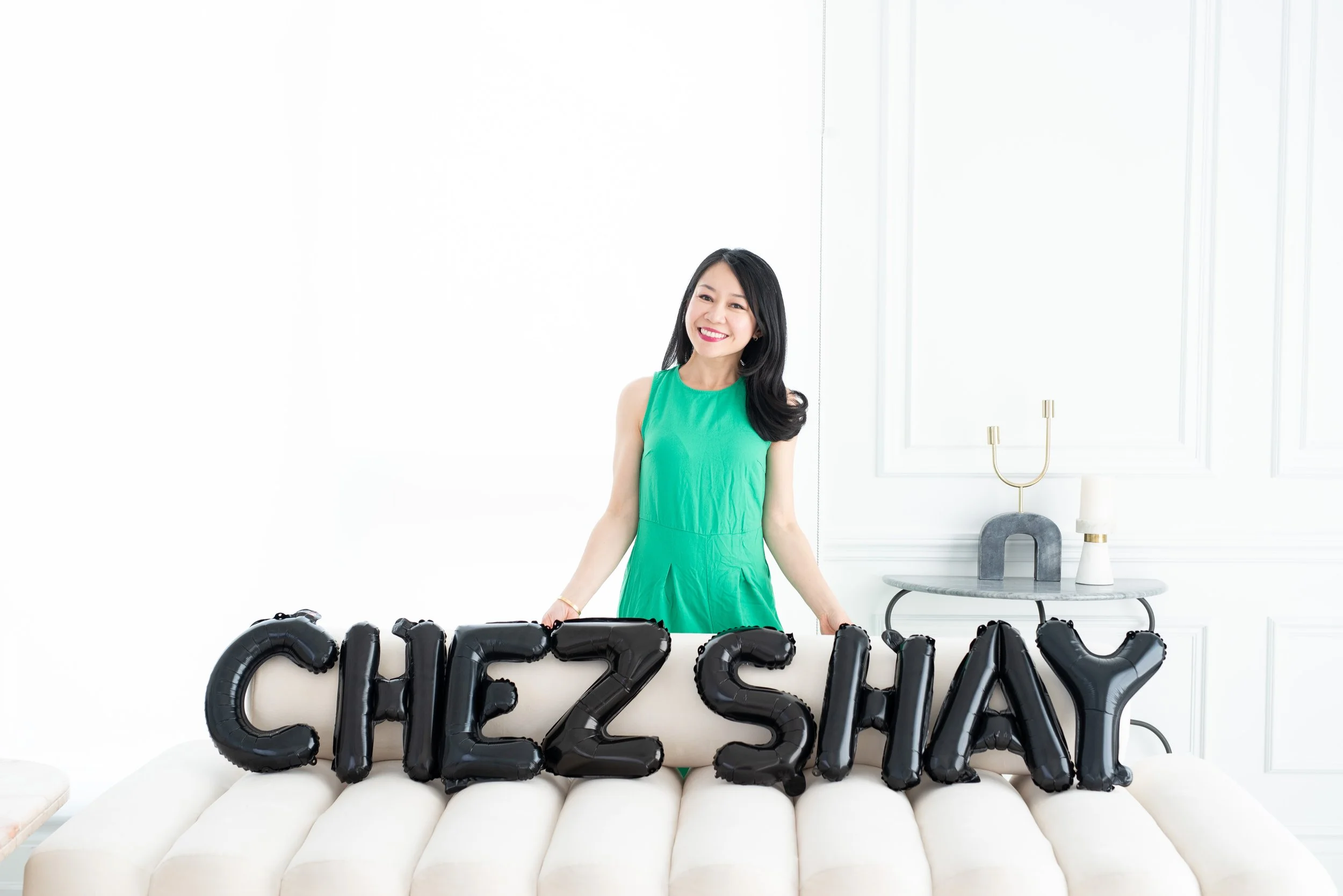 A woman in a green dress smiling and standing behind black balloon letters spelling "CHEZ SHAY" on a white sofa in a bright, minimalistic room.