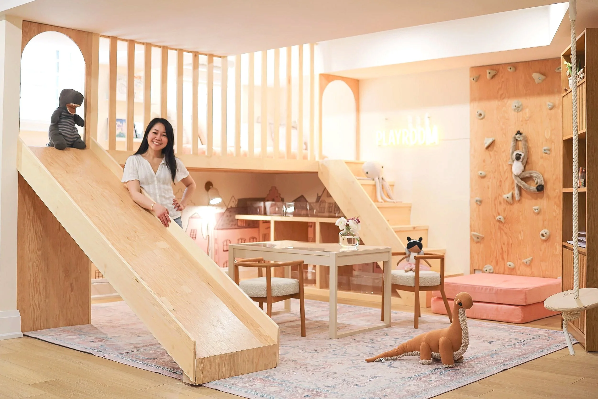 A woman smiling in a playroom with a wooden slide, climbing wall, stuffed animals, and a small table with chairs.